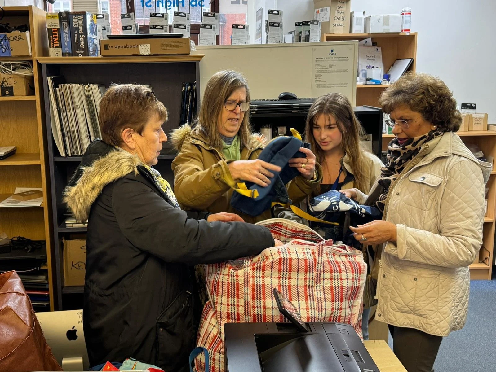 Four women standing around a table in an office or store, looking at and handling bags and items, with shelves filled with boxes, books, and office supplies in the background.
