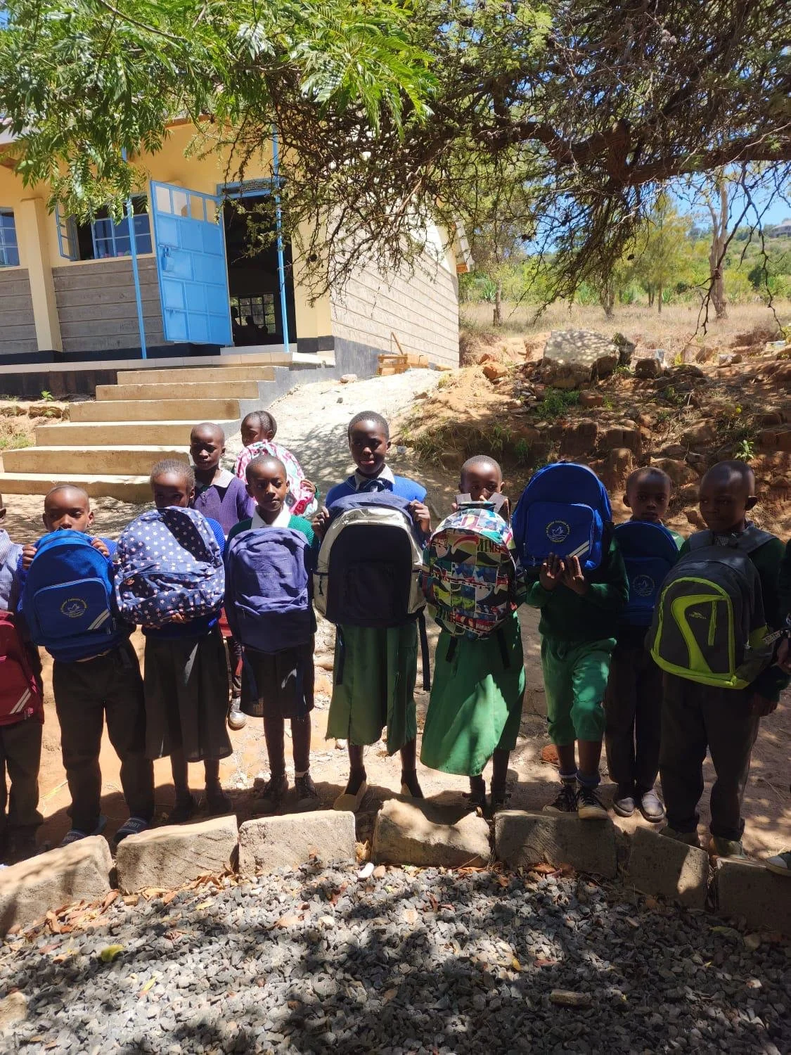 Group of school children with backpacks standing outdoors in front of a building with open blue doors, under the shade of a tree.