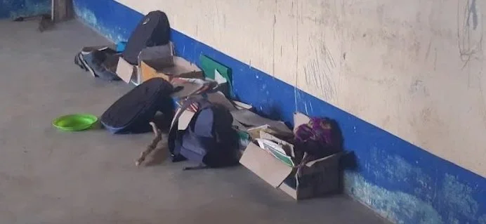 A backpack, books, and boxes of supplies are scattered on the floor near a wall in what appears to be a public space.