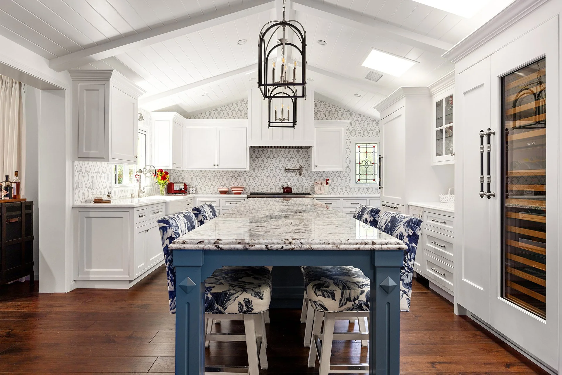 Spacious kitchen with vaulted white ceilings, patterned backsplash, a large blue island with a stone countertop, and floral upholstered stools set on warm wood floors.