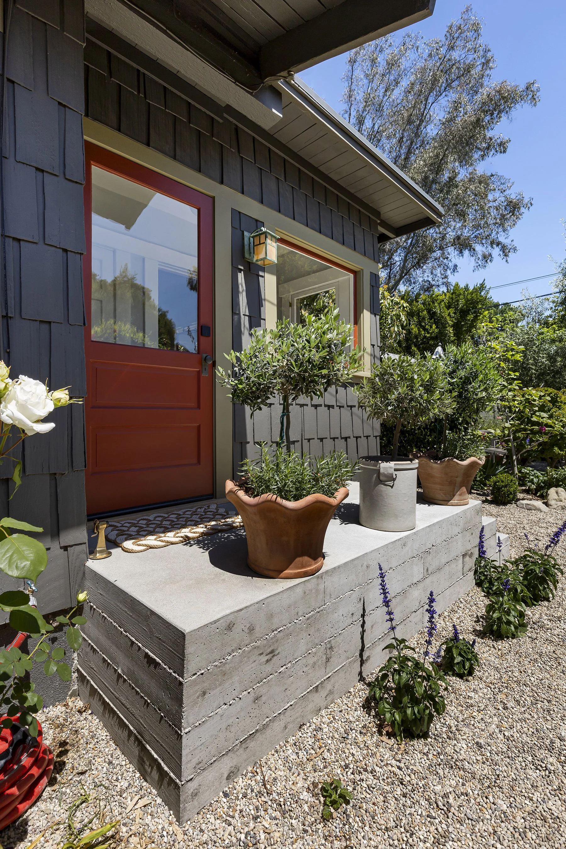 Front porch with potted plants in decorative pots, a red door, and a window, surrounded by a garden with purple flowers and gravel ground, under a clear blue sky.