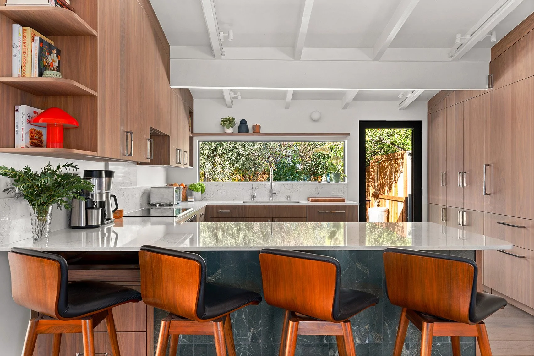 Modern kitchen with warm wood cabinets, a marble island, mid-century barstools, and a wide window overlooking greenery.