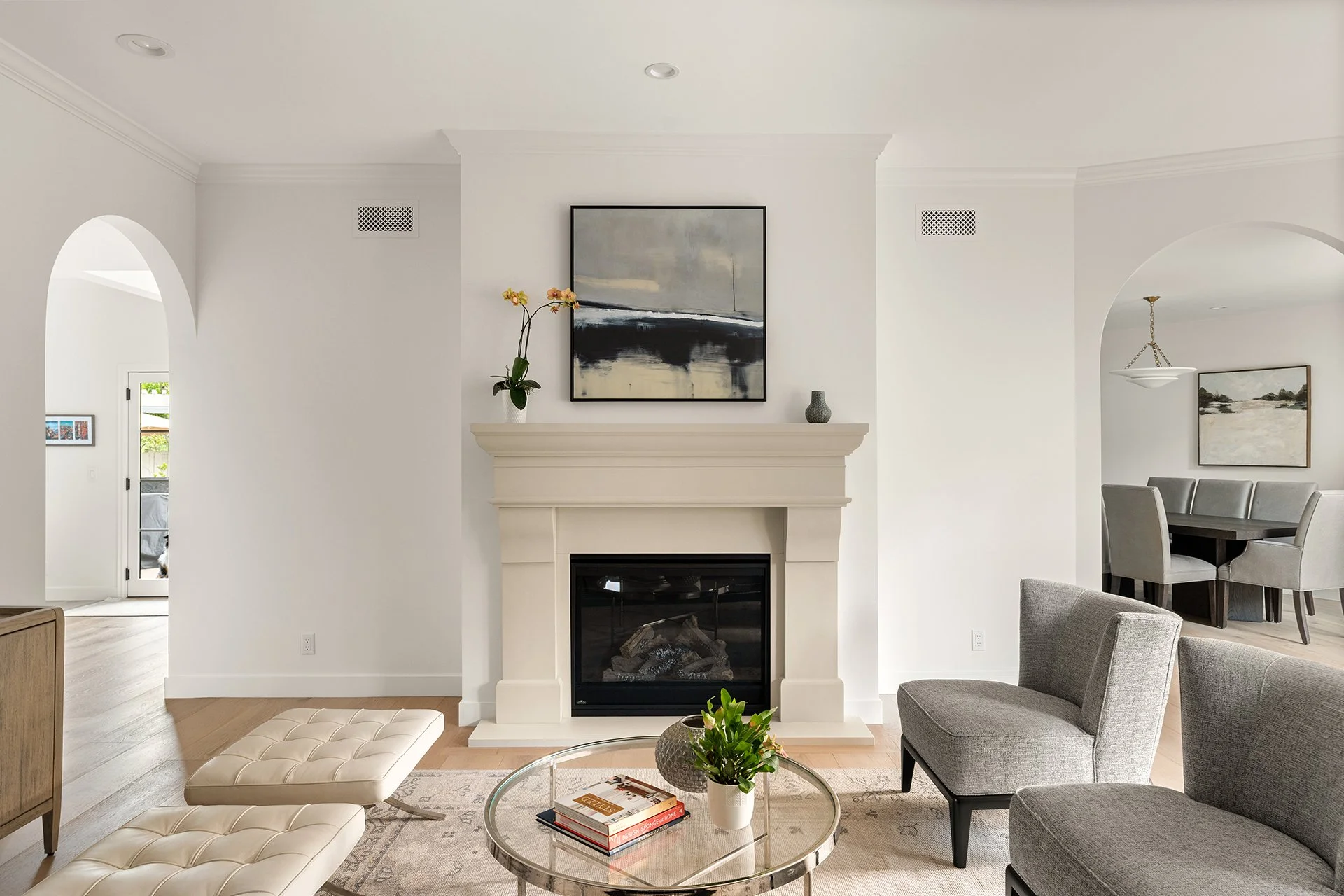 Living room in La Cañada custom home remodel featuring white fireplace mantel, abstract artwork, modern gray chairs, and glass coffee table styled with books and greenery, with open dining area beyond.
