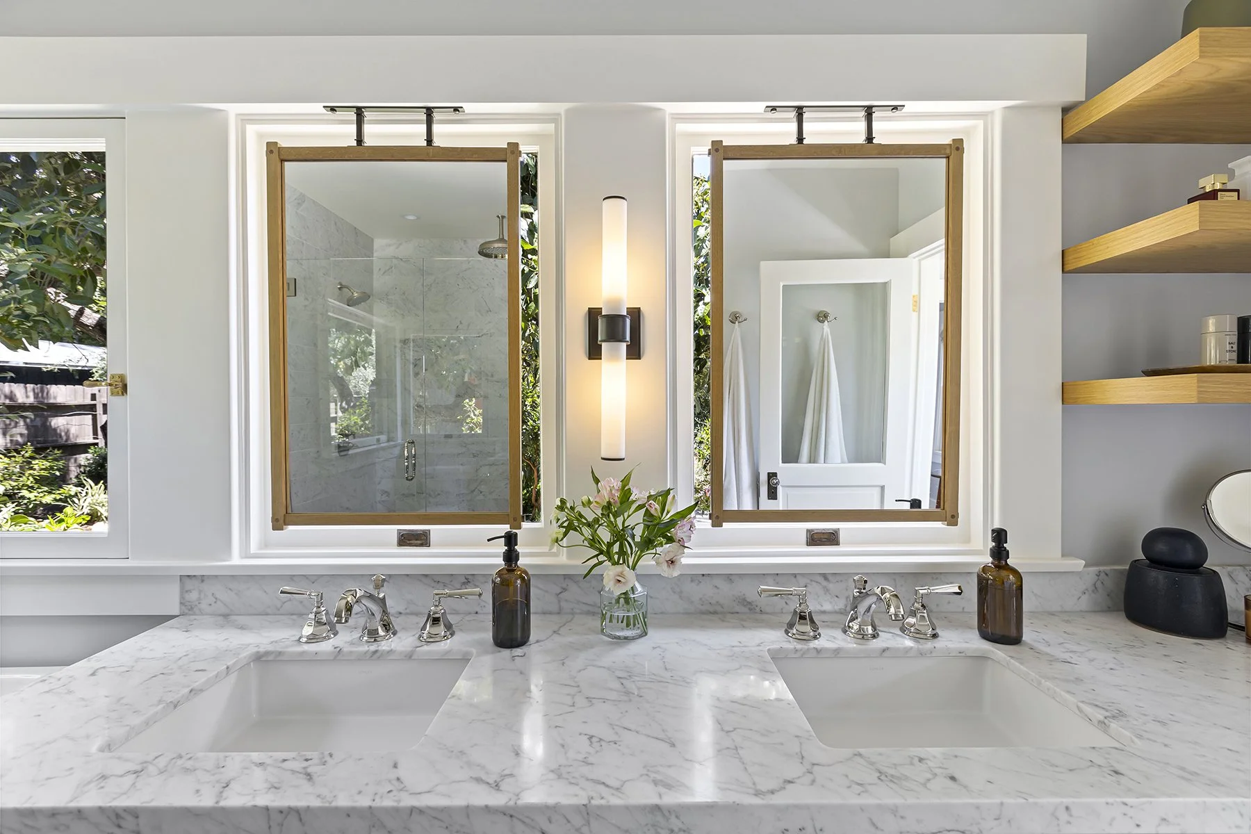 Double-sink marble vanity with chrome fixtures, warm wood-framed mirrors suspended in front of bright windows, a central wall sconce, and open shelves styled with modern accessories.