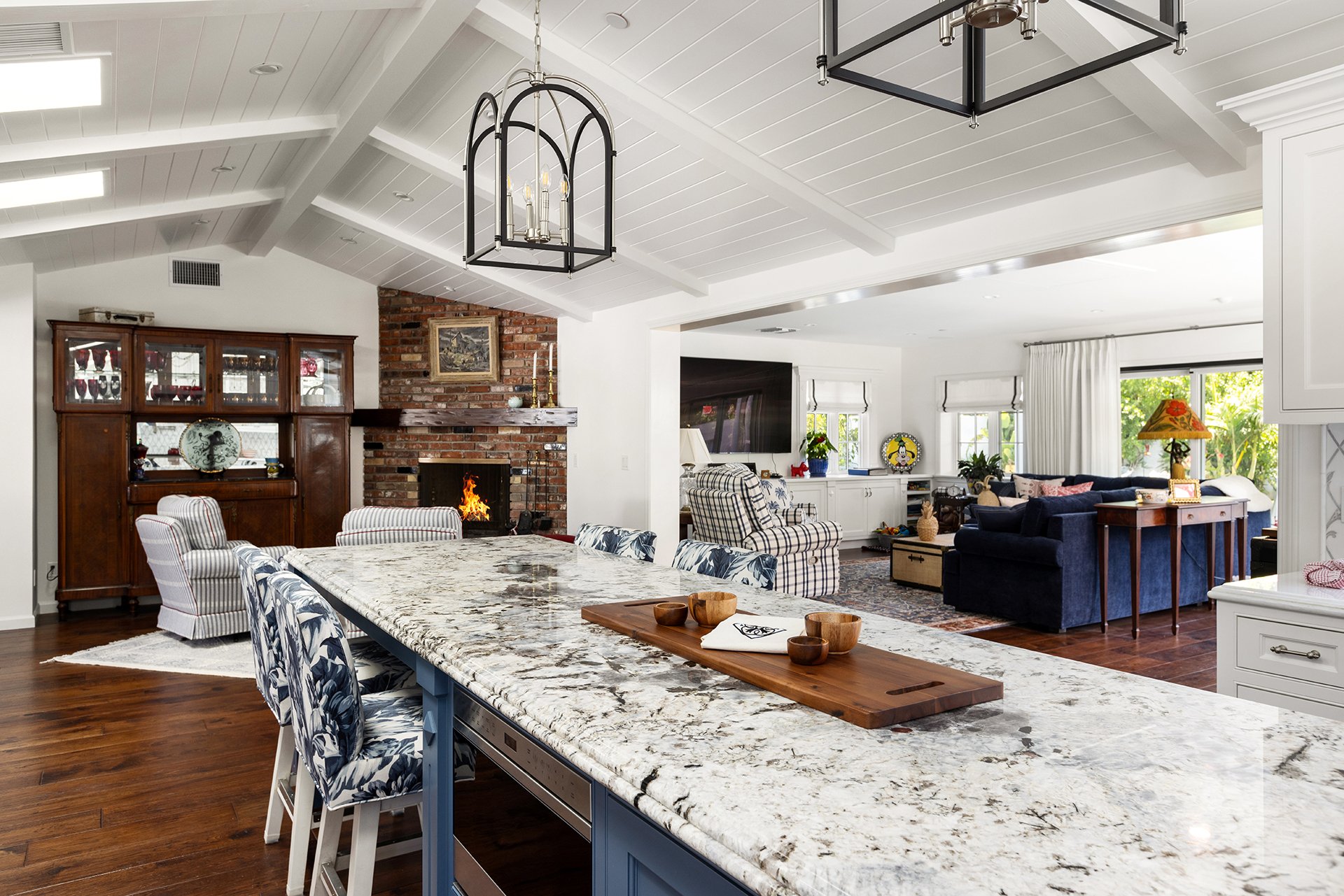 Open-concept kitchen and living room with vaulted white ceilings, marble island, brick fireplace, and traditional furnishings.