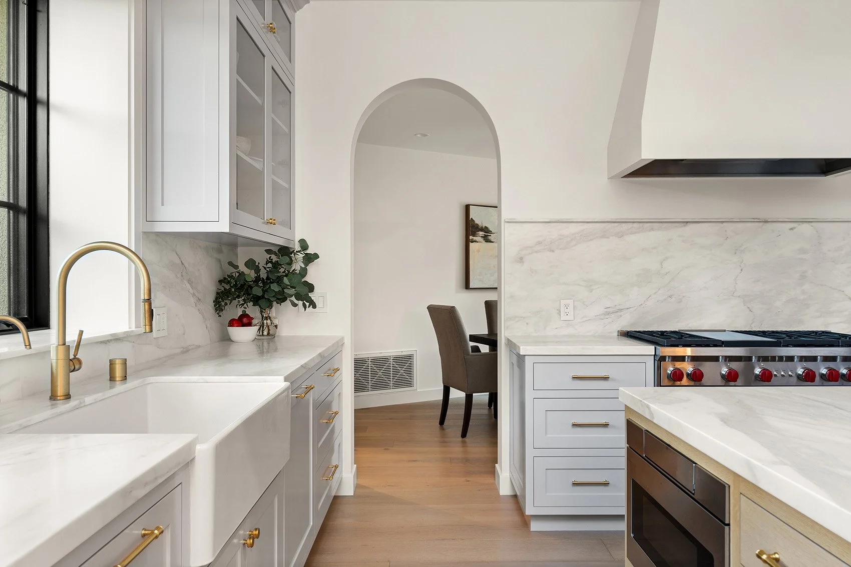 Bright, modern kitchen with light gray cabinets, brass hardware, a white farmhouse sink, marble countertops and backsplash, and a black-framed window overlooking green foliage.