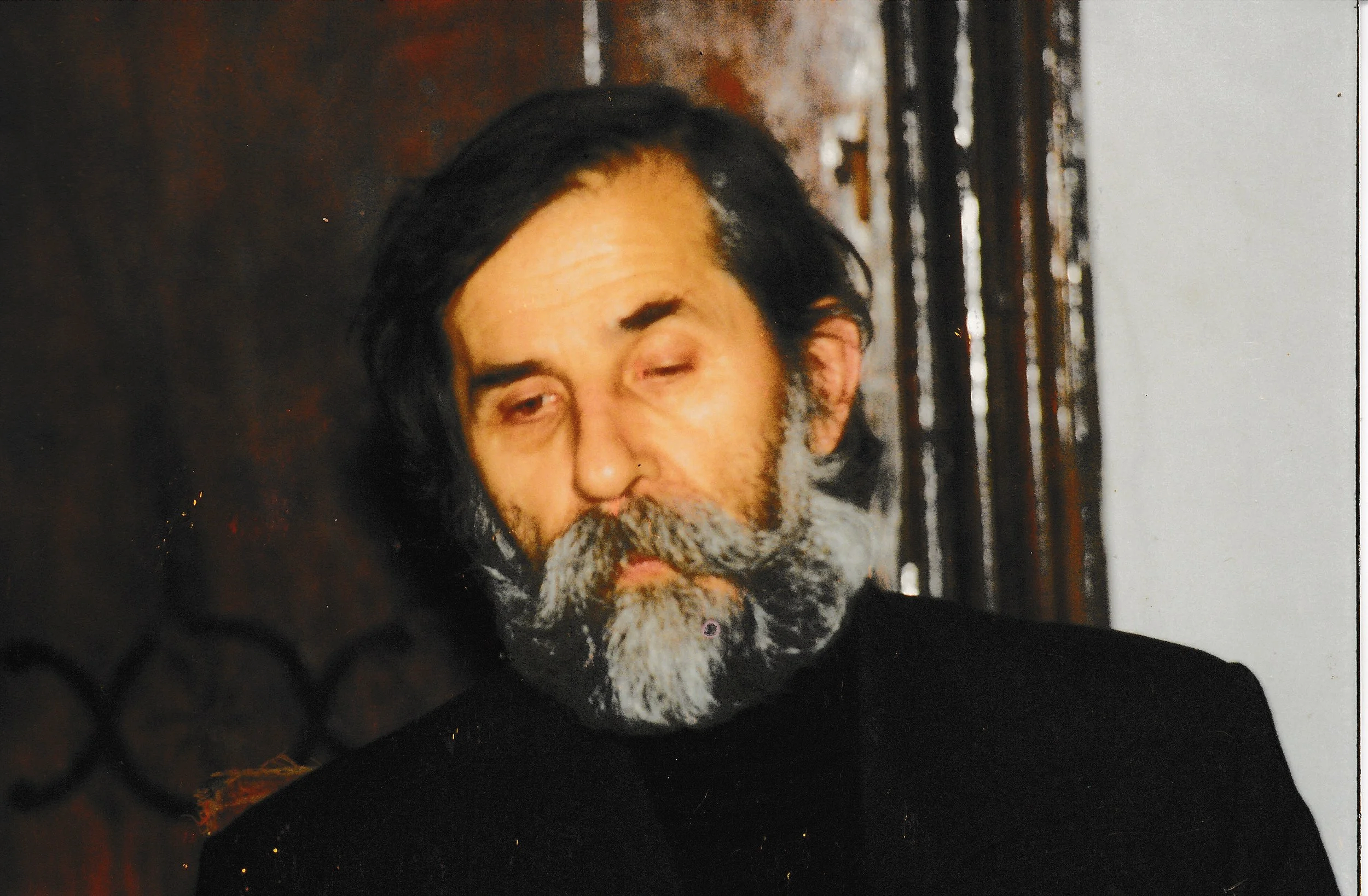 A man with a beard and mustache, with one eye closed and the other partially open, wearing a black shirt, standing in front of a dark and plain background with some wooden elements.
