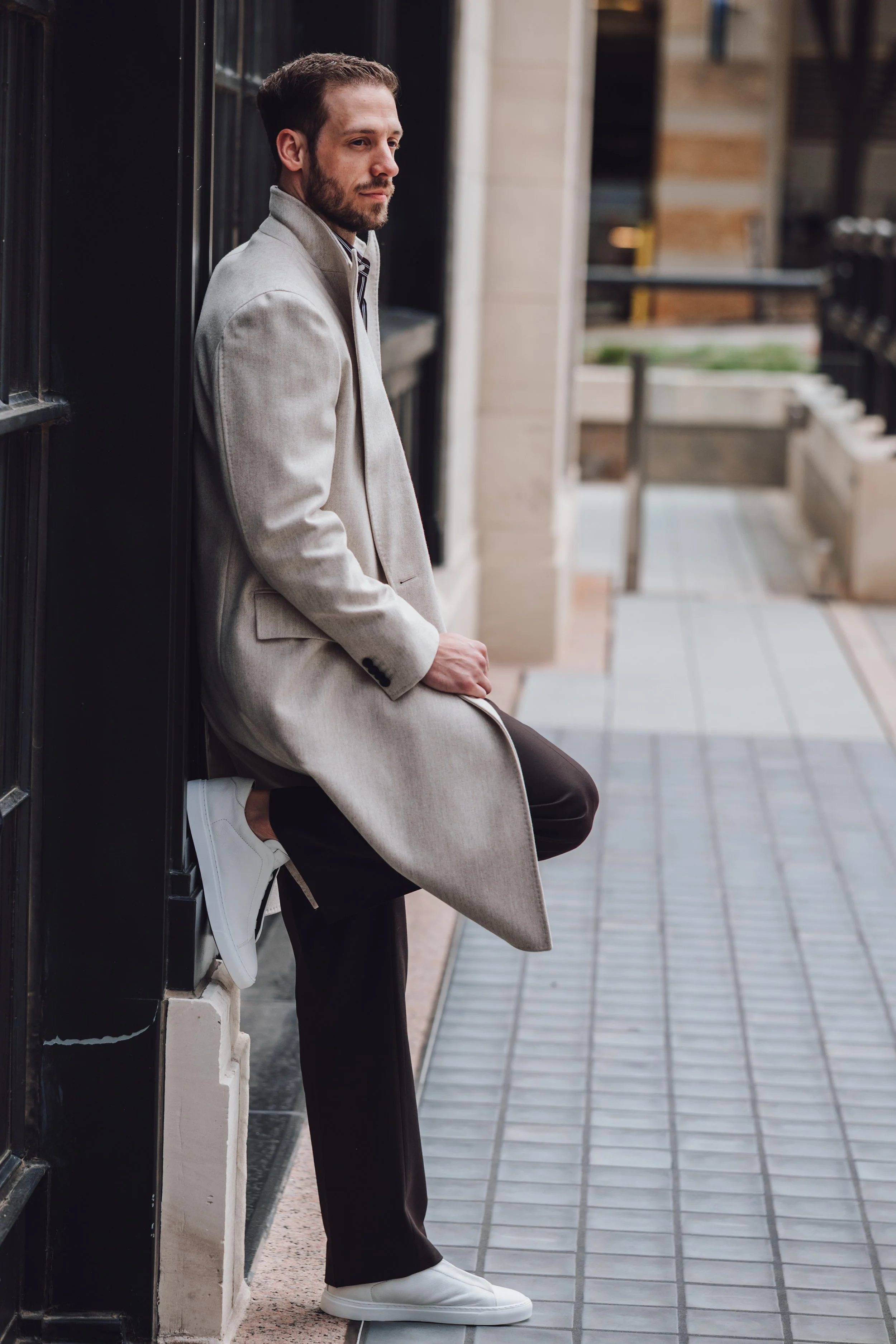 A man in a beige coat, black pants, and white sneakers is sitting against a black wall in an urban setting, with a thoughtful expression.