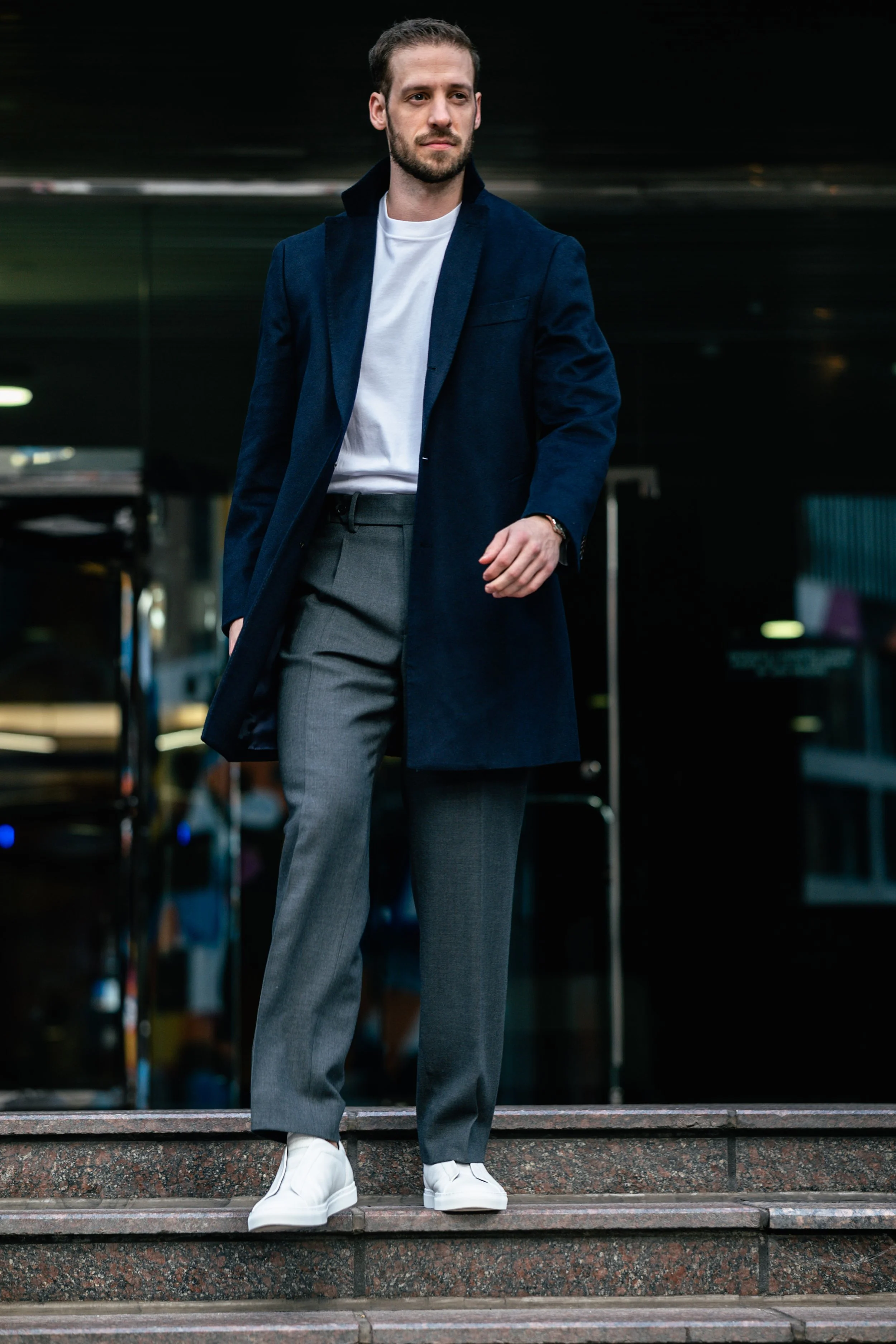 A man dressed in a white t-shirt, navy coat, and gray pants walking down steps outside a building at night.