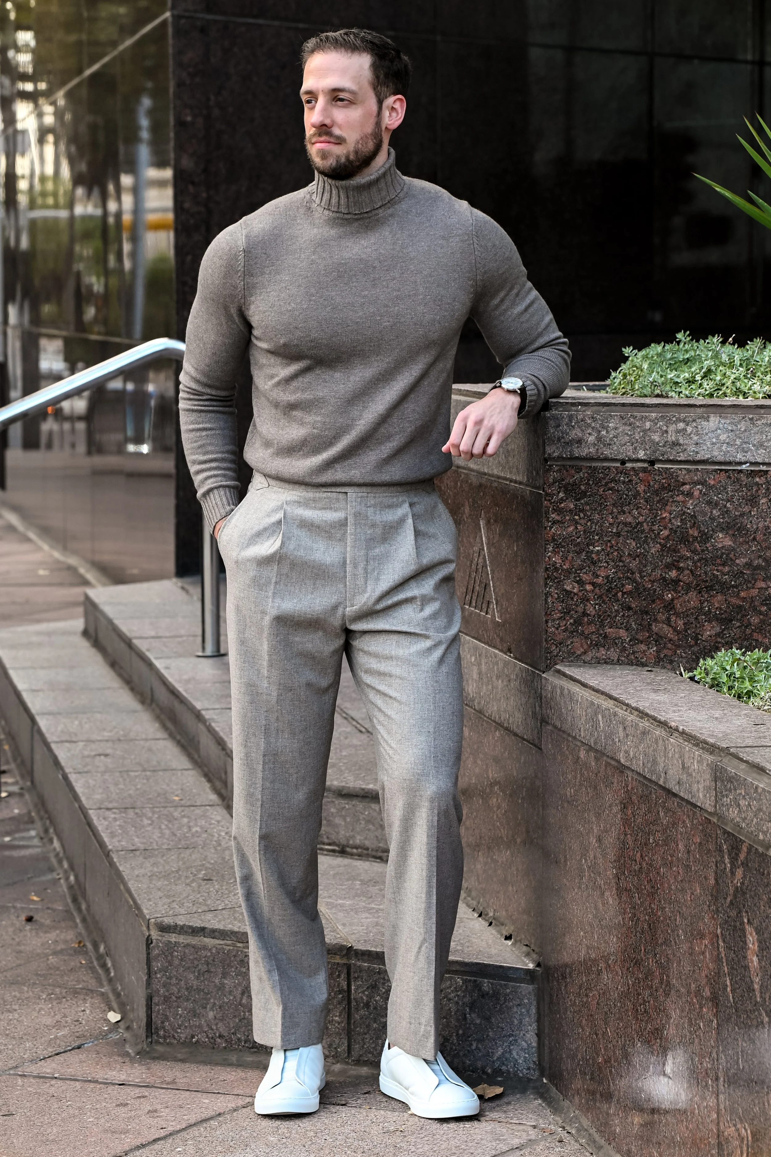 A man in a gray turtleneck sweater and light gray dress pants stands outside beside a stone and bronze planters, wearing white sneakers and a watch, with his hand resting on the edge of the planter against a modern building.