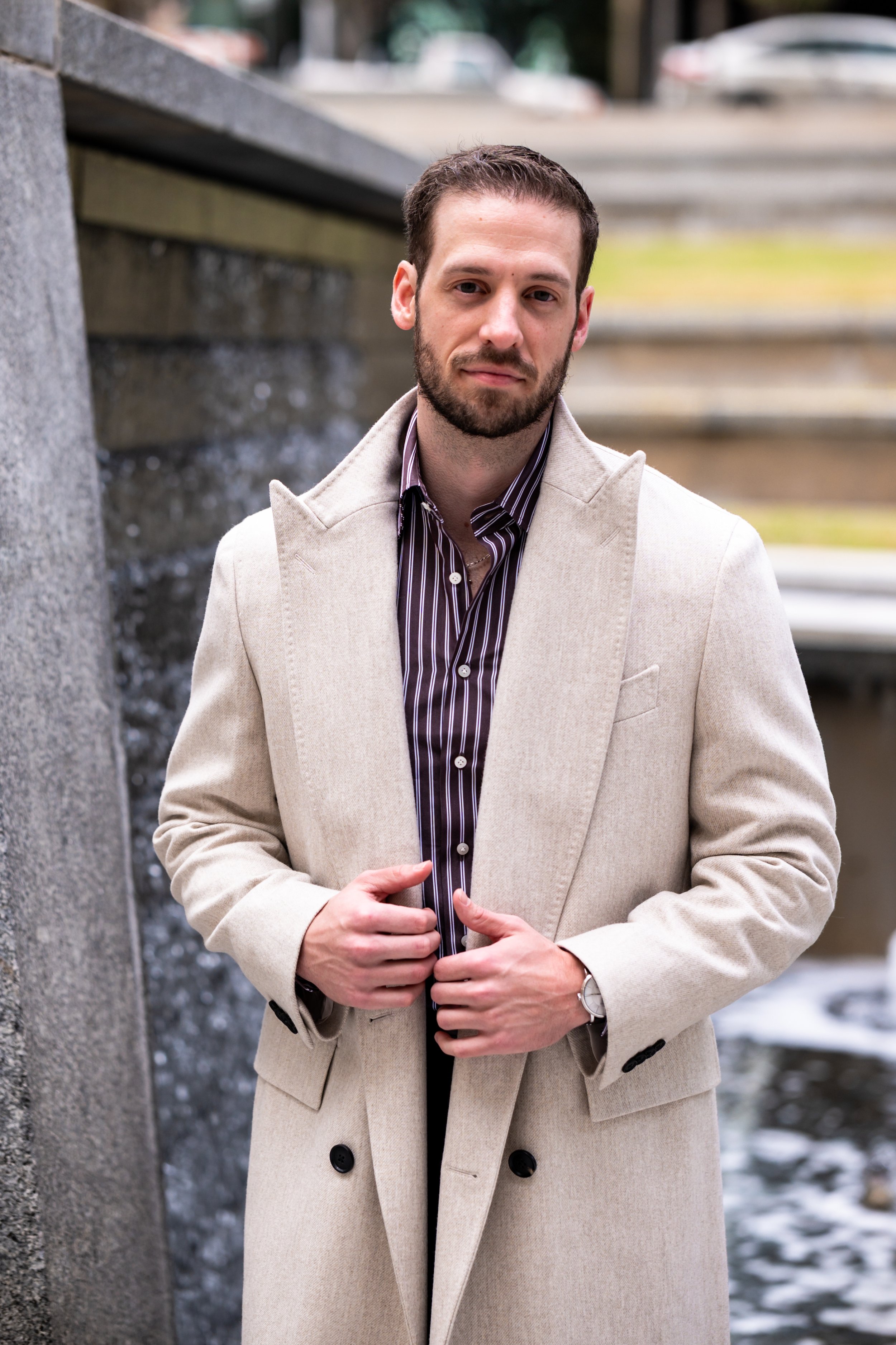 A man in a light beige coat and a purple and white striped shirt standing outdoors in front of a stone wall with water streaming down it.
