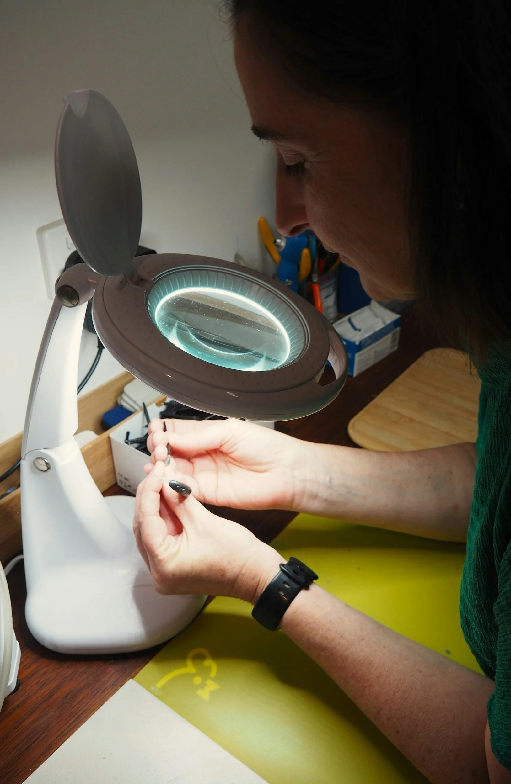 An audiologist at Integrity Hearing Services in Lakewood, Washington, sitting at a desk using a lamp to look closely at the inside of a hearing aid and repair the hearing aid.