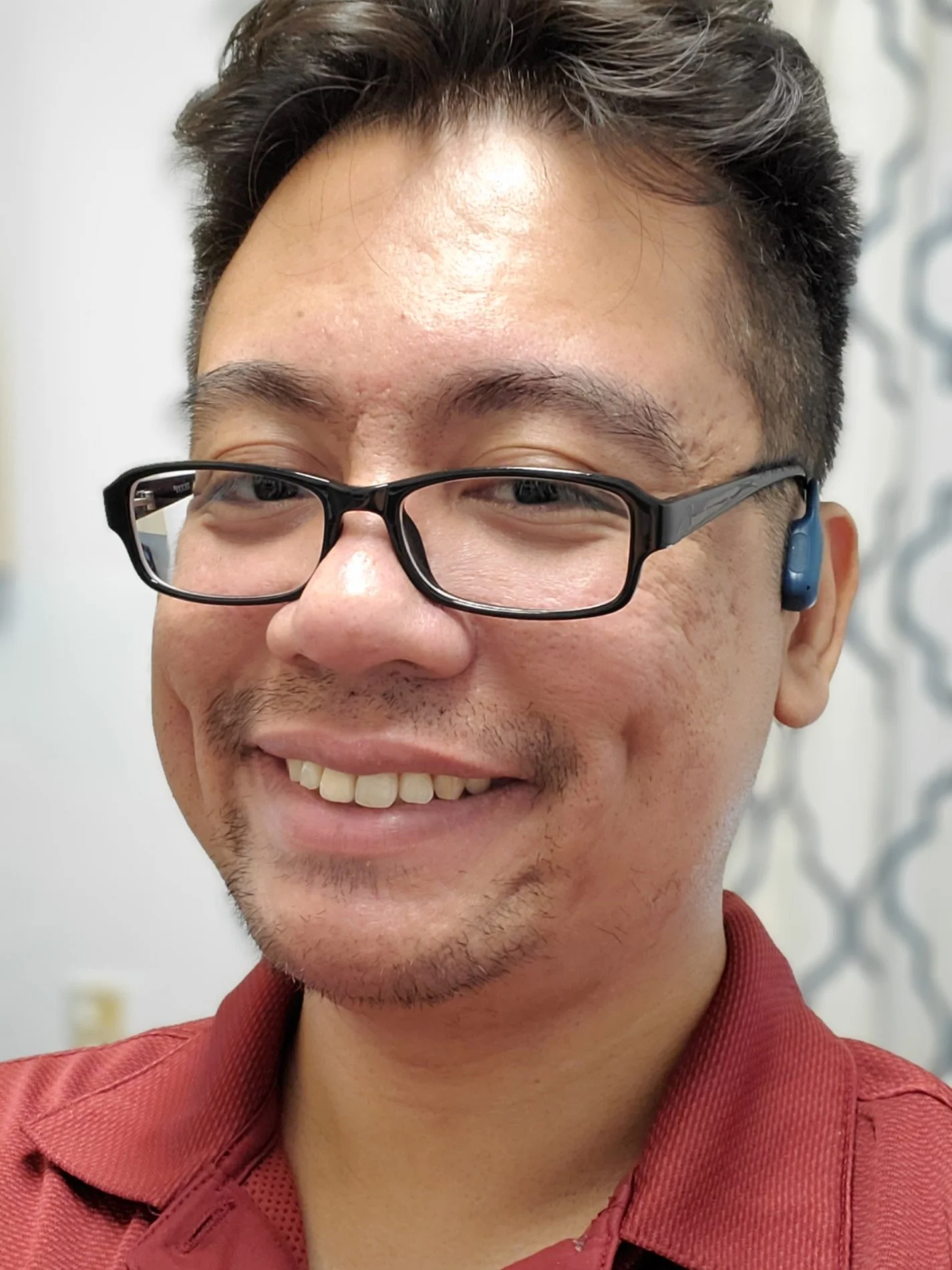 A smiling man wearing glasses and a red shirt, with a hearing aid behind his ear, standing indoors near a patterned curtain.