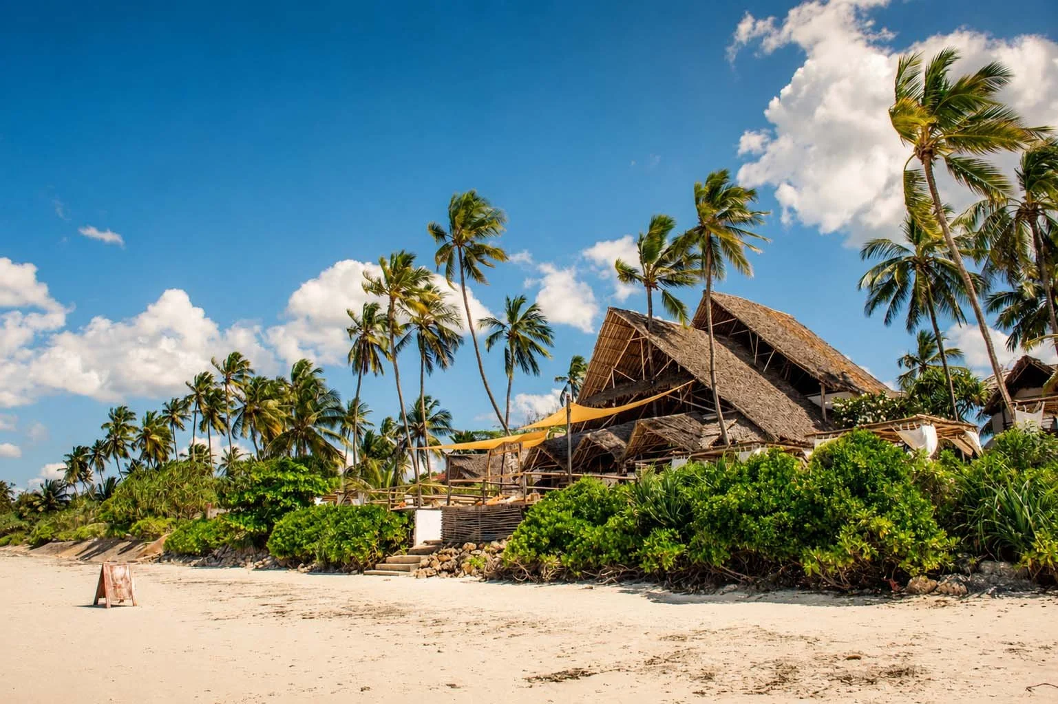 A tropical beach with white sand, lush green bushes, tall palm trees, and rustic thatched-roof huts under a blue sky with scattered white clouds.