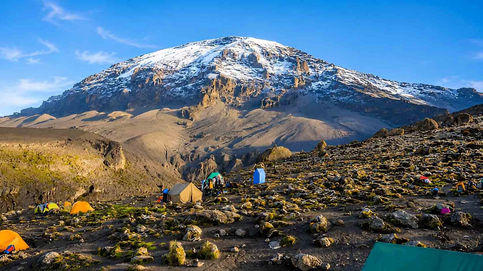 Campground with tents set up on rocky terrain at the base of a snow-capped mountain under a clear blue sky.