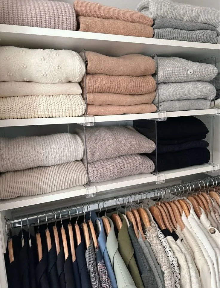 Closet shelf with folded sweaters in neutral colors and a clothing rack with hangers holding various shirts and jackets.