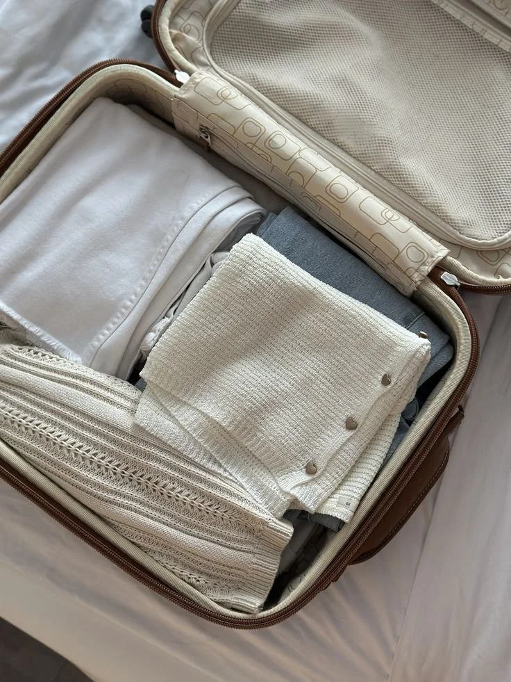 Open suitcase packed with laundry, including white and gray clothing, and a folded beige towel or cloth.