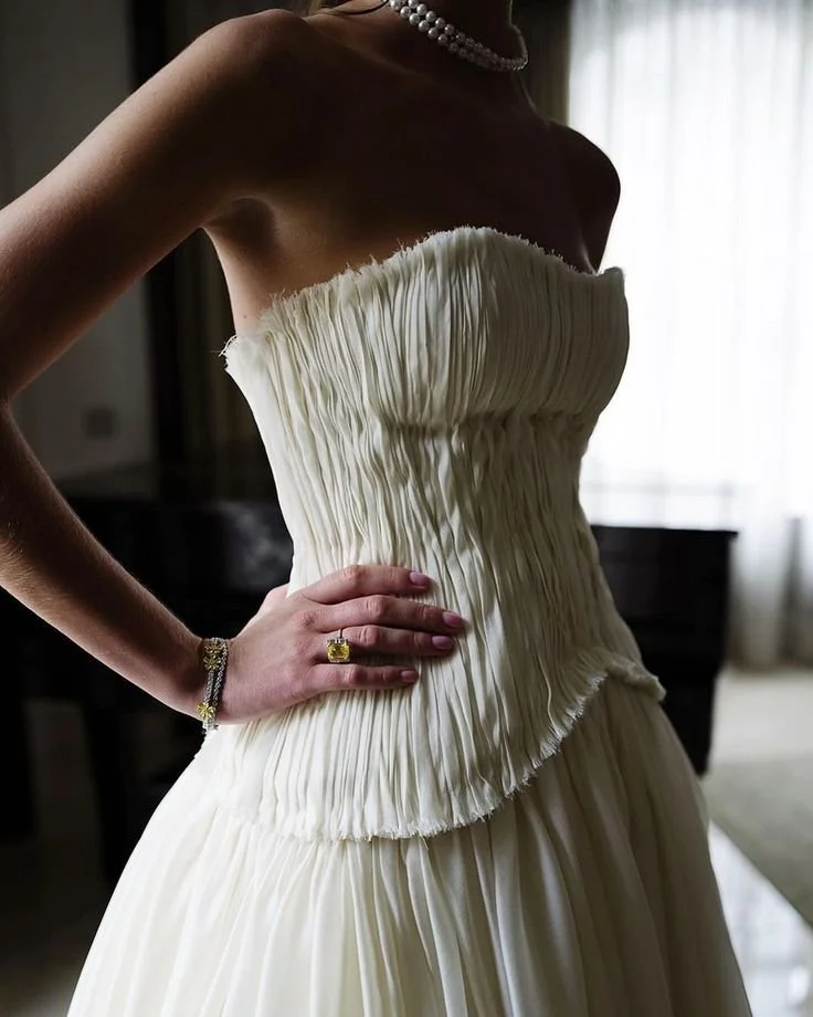 Close-up of a woman in a strapless, ruched wedding gown, wearing a pearl necklace and bracelet, and a ring with a yellow gemstone, standing indoors with a curtain in the background.