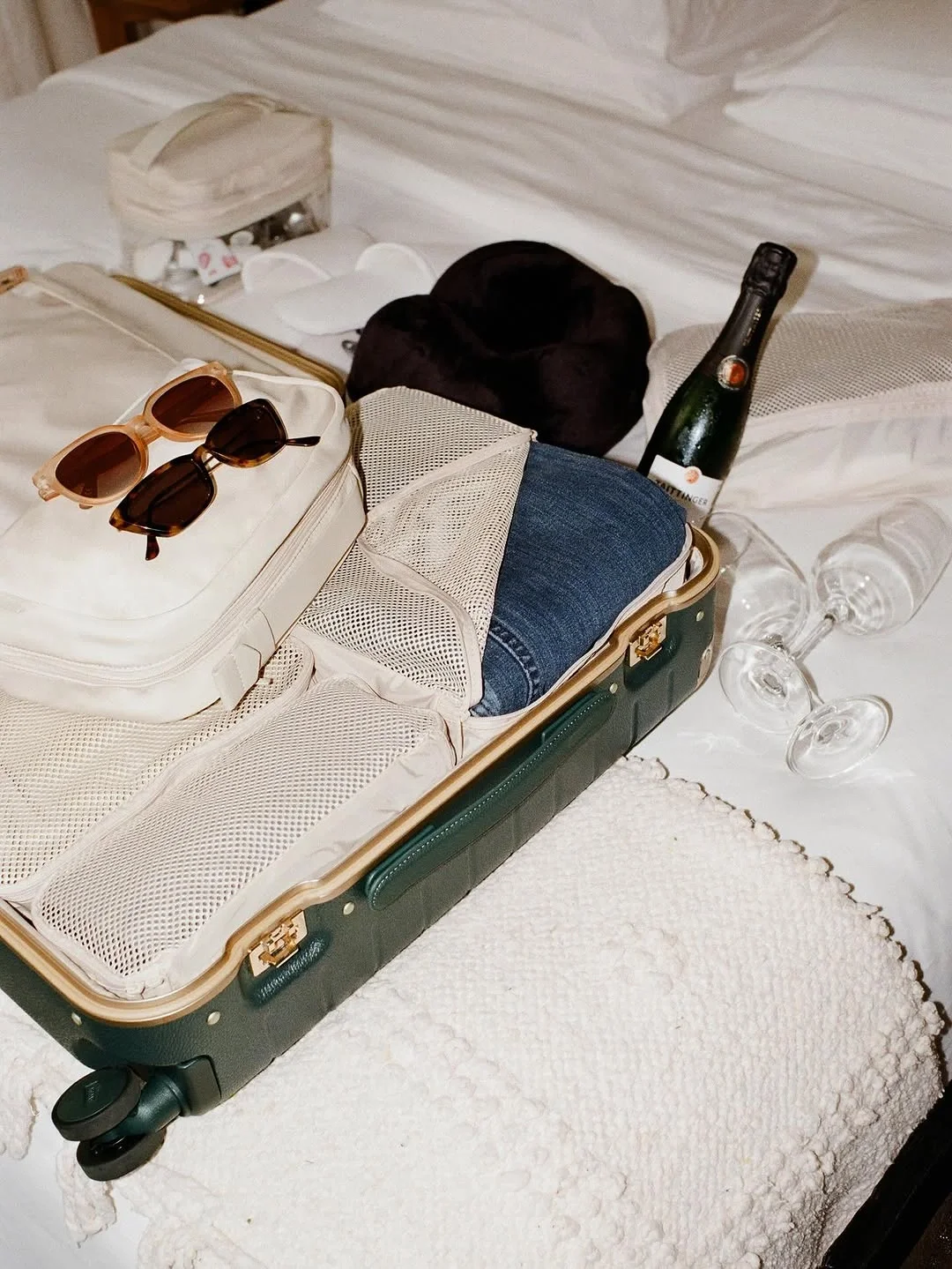 Open suitcase with sunglasses, clothes, and travel items on a bed, with a bottle and glasses nearby.