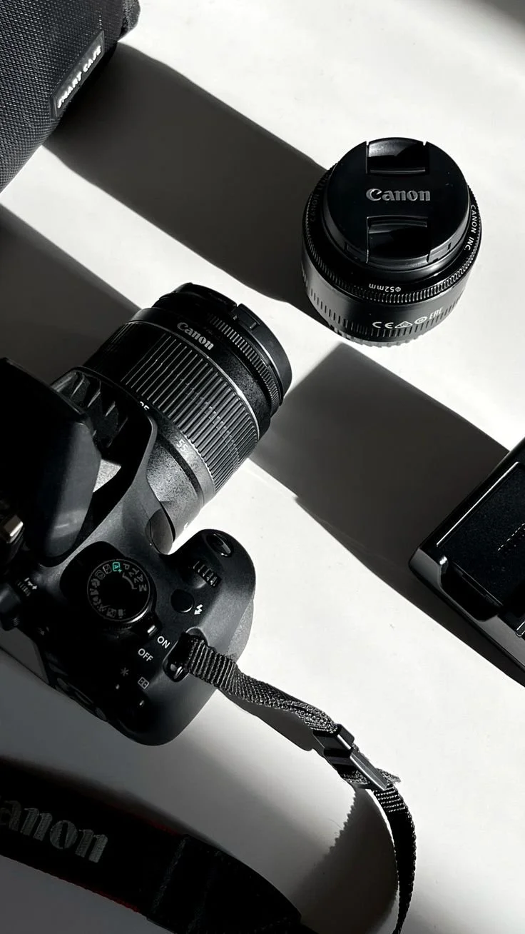 A Canon camera with a detachable lens, a camera body cap, and a camera strap on a white surface with strong shadows.