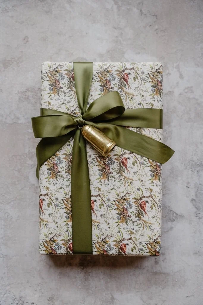 A wrapped gift box with floral patterned paper, tied with a wide green satin ribbon and decorated with a small gold ornament.