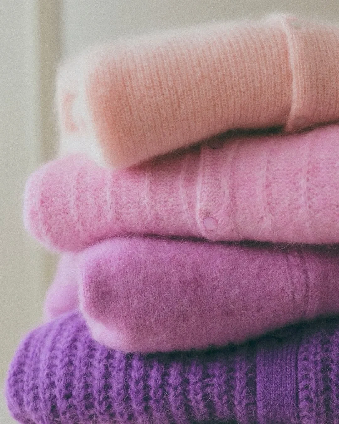 Stacked colorful knitted sweaters in beige, pink, lavender, and purple.