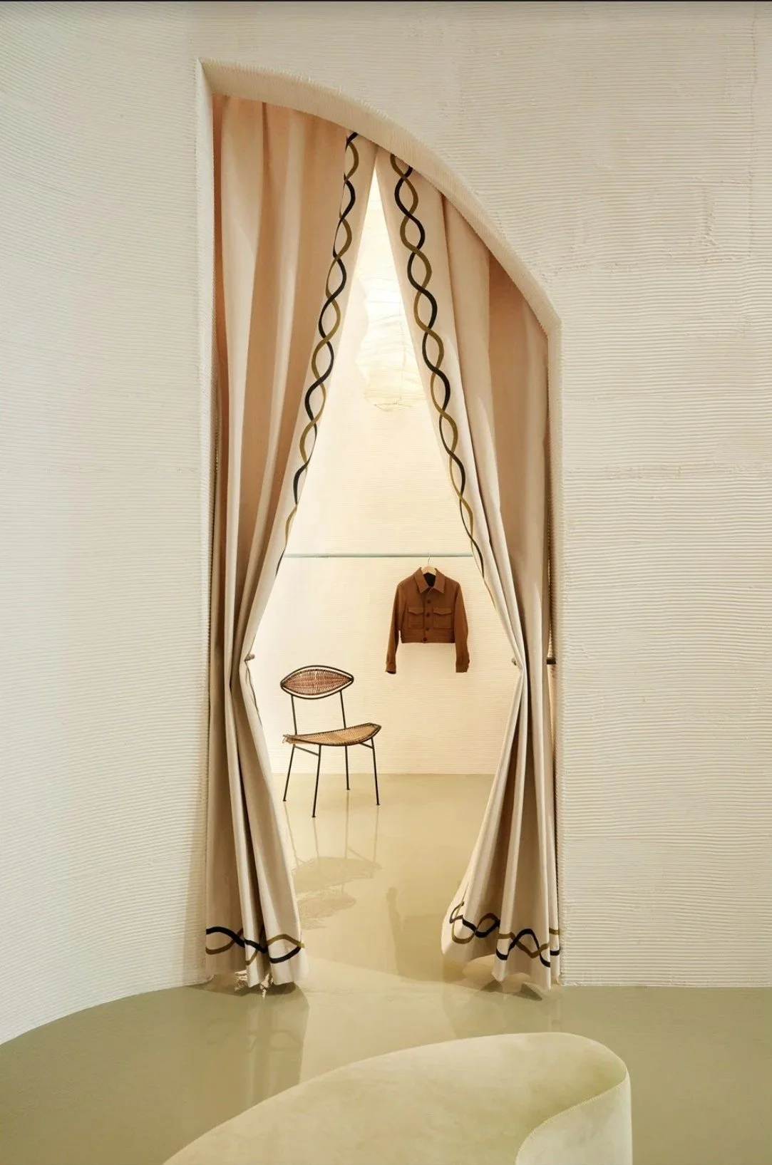 An ornate doorway with cream-colored curtains that have black and gold woven trim. Behind the curtains is a minimalist room with a chair and a brown jacket hanging on the wall.