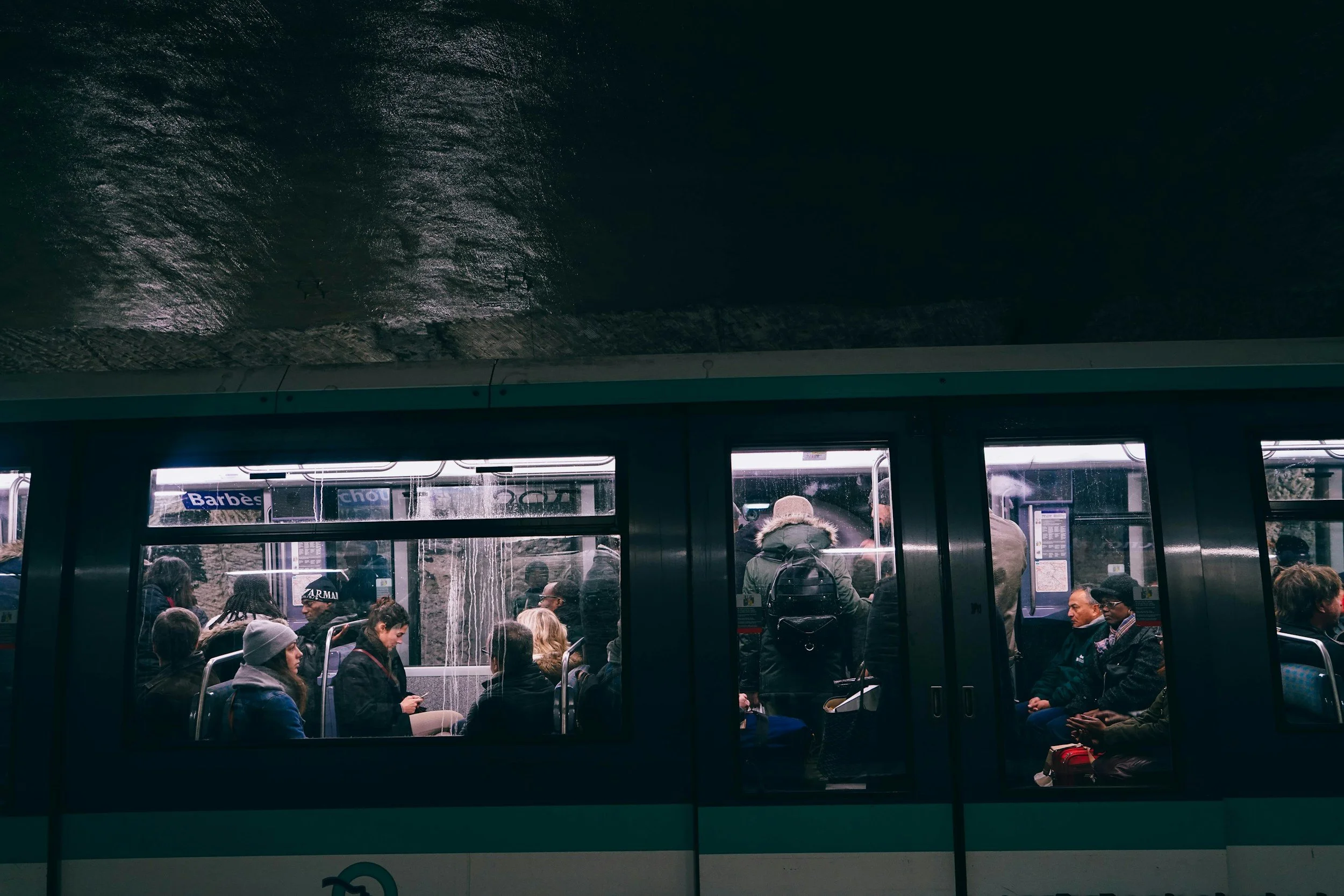 View of a crowded subway train interior through the glass doors. Passengers are seated and standing, with some using phones. Overhead signs and station advertisements are visible through the window.