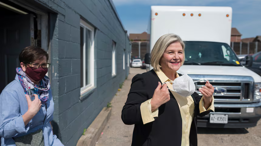 Two women standing outside near a gray building and a white delivery truck. The woman in the foreground is wearing a black blazer and holding a face mask, smiling. The woman in the background is wearing glasses, a red face mask, and a blue shirt with a colorful scarf, giving a thumbs-up.