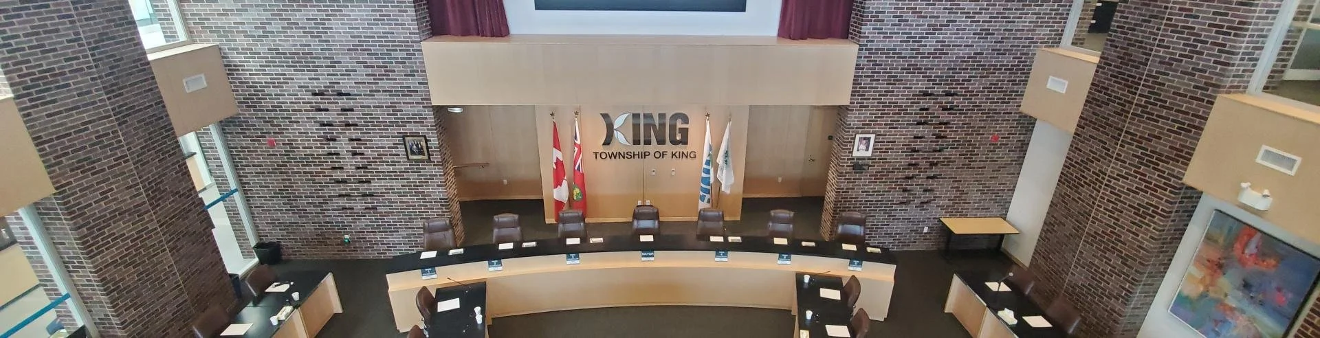 Empty city council chamber with a curved desk, chairs, flags, and a large wall sign reading "King Township of King."