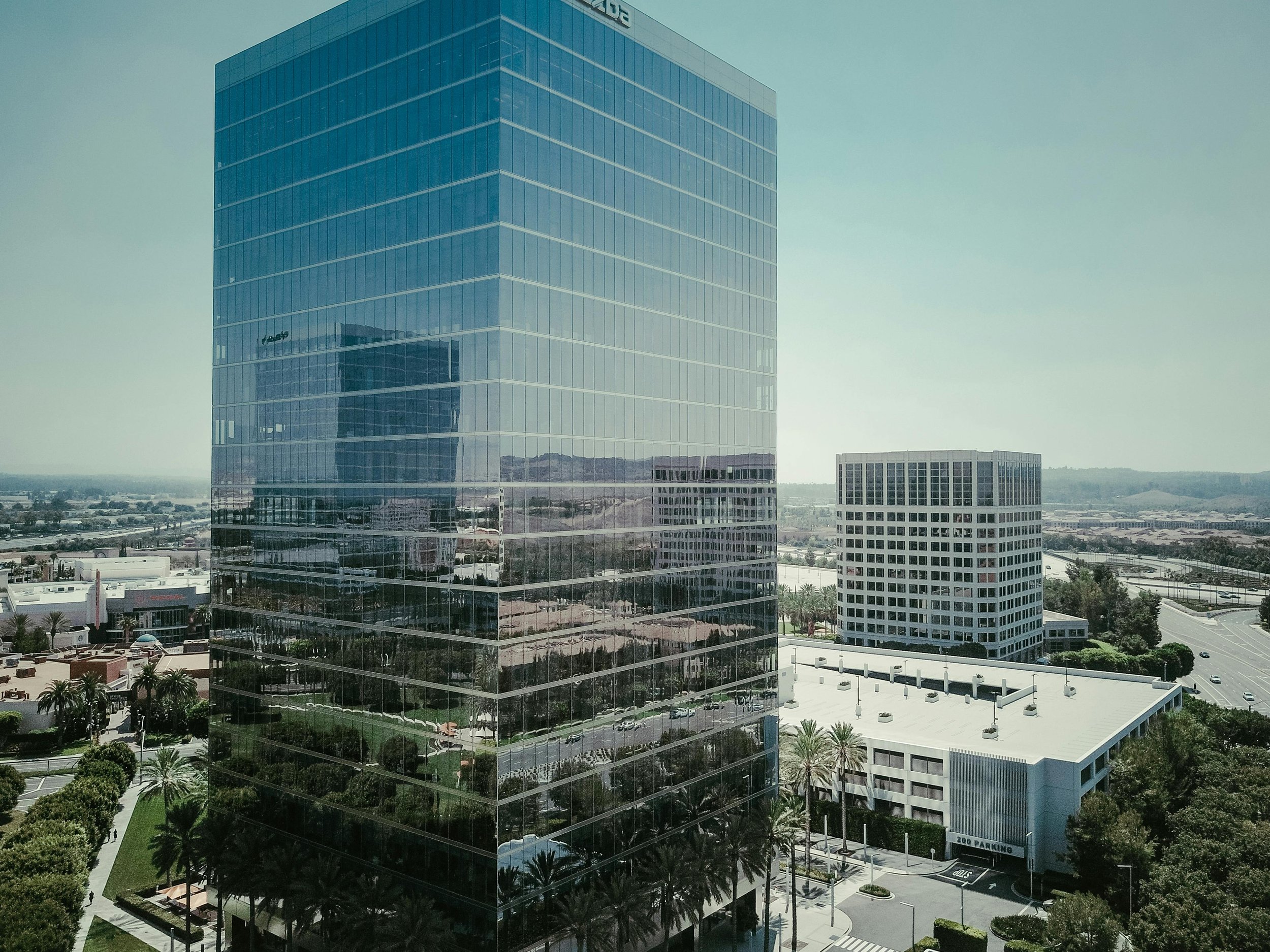 A tall glass office building reflecting the surrounding cityscape, with smaller office buildings, parking lots, roads, and trees in the background under a clear sky.