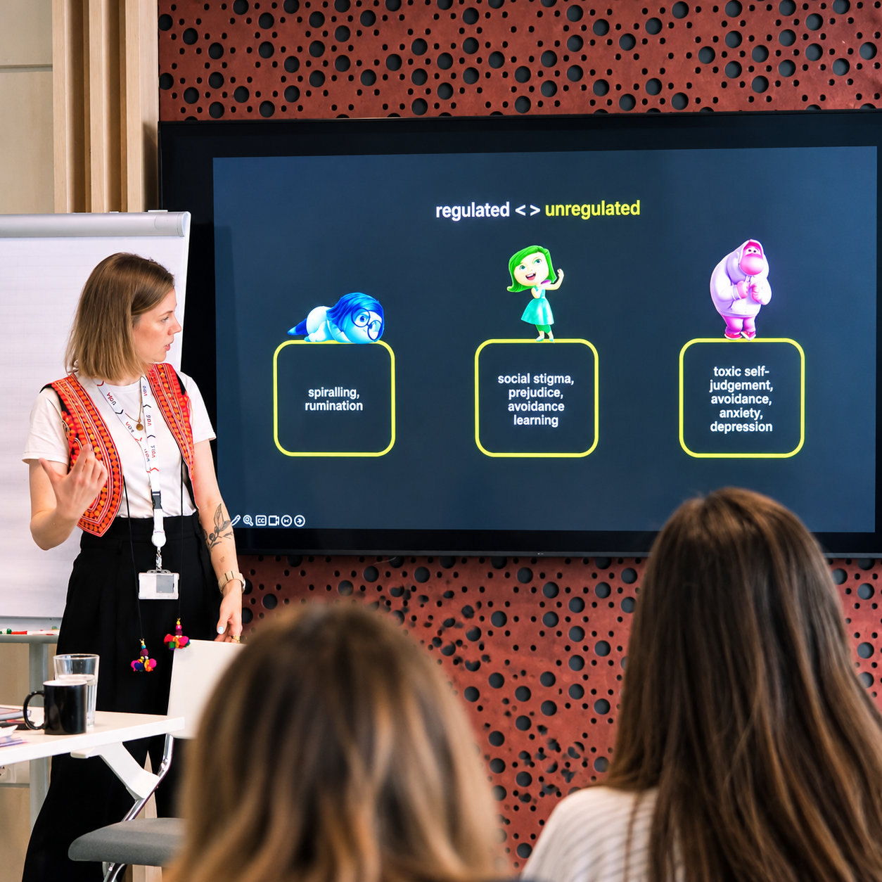 A woman giving a presentation on regulation of behaviors, showing three cartoon characters with explanations: a blue character laying down labeled 'spiraling, rumination,' a green character standing with a raised hand labeled 'social stigma, prejudice, avoidance learning,' and a pink character standing with crossed arms labeled 'toxic self-judgement, avoidance, anxiety, depression.'