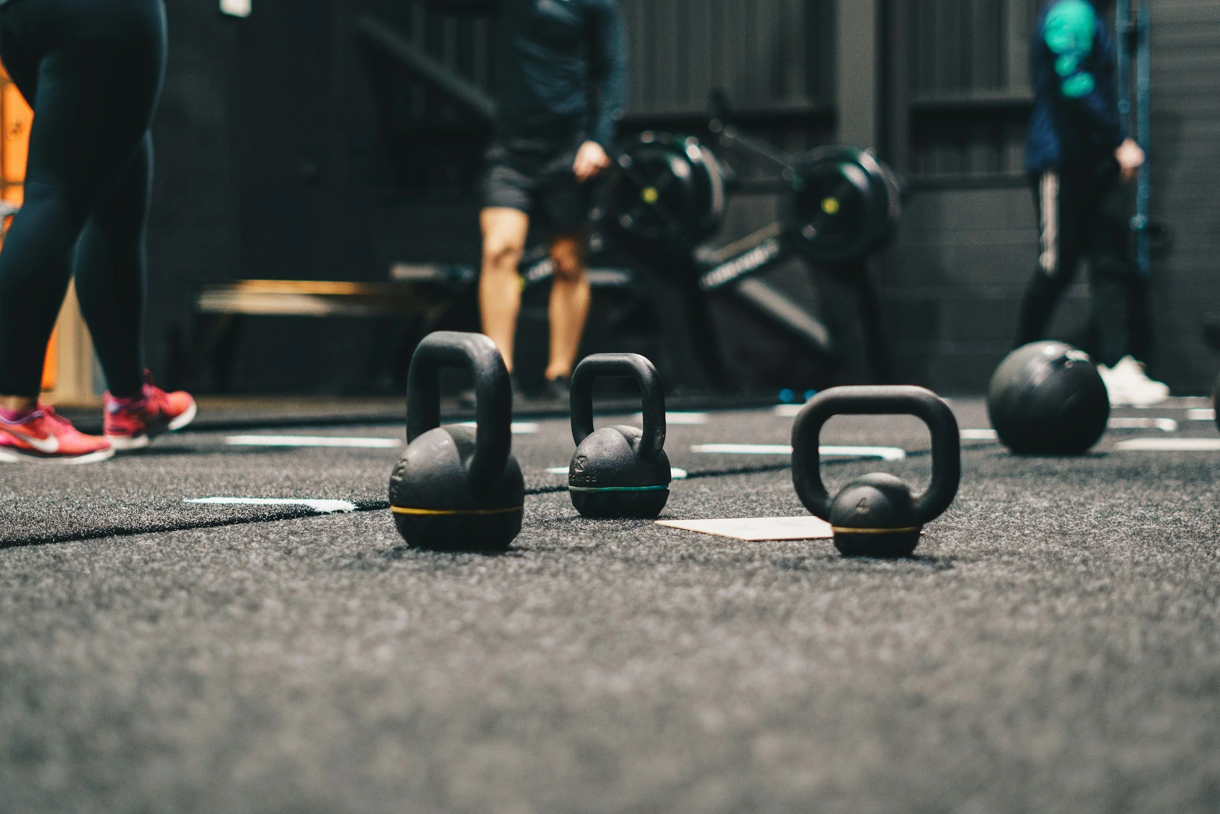 Kettlebells and black medicine ball on black gym floor with people working out in the background.