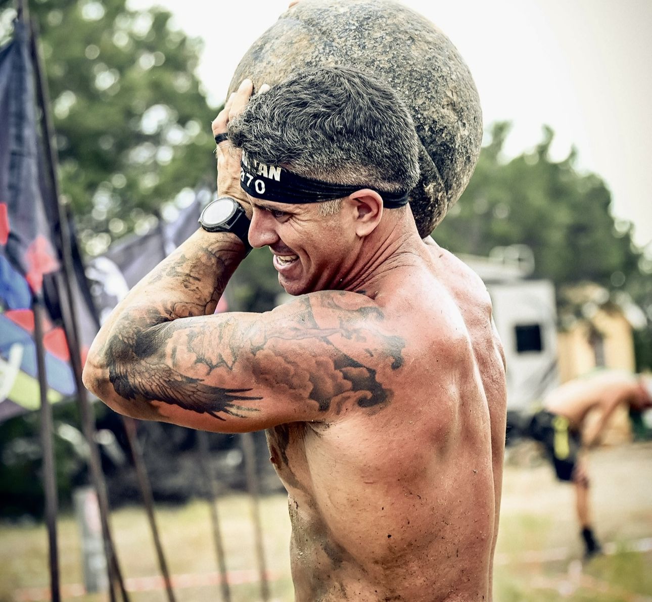 A muscular man (Ryan Padilla, Apogee Fitness Training, Spartan Race) is participating in a tough fitness event, carrying a large rock on his shoulder during an outdoor obstacle course.