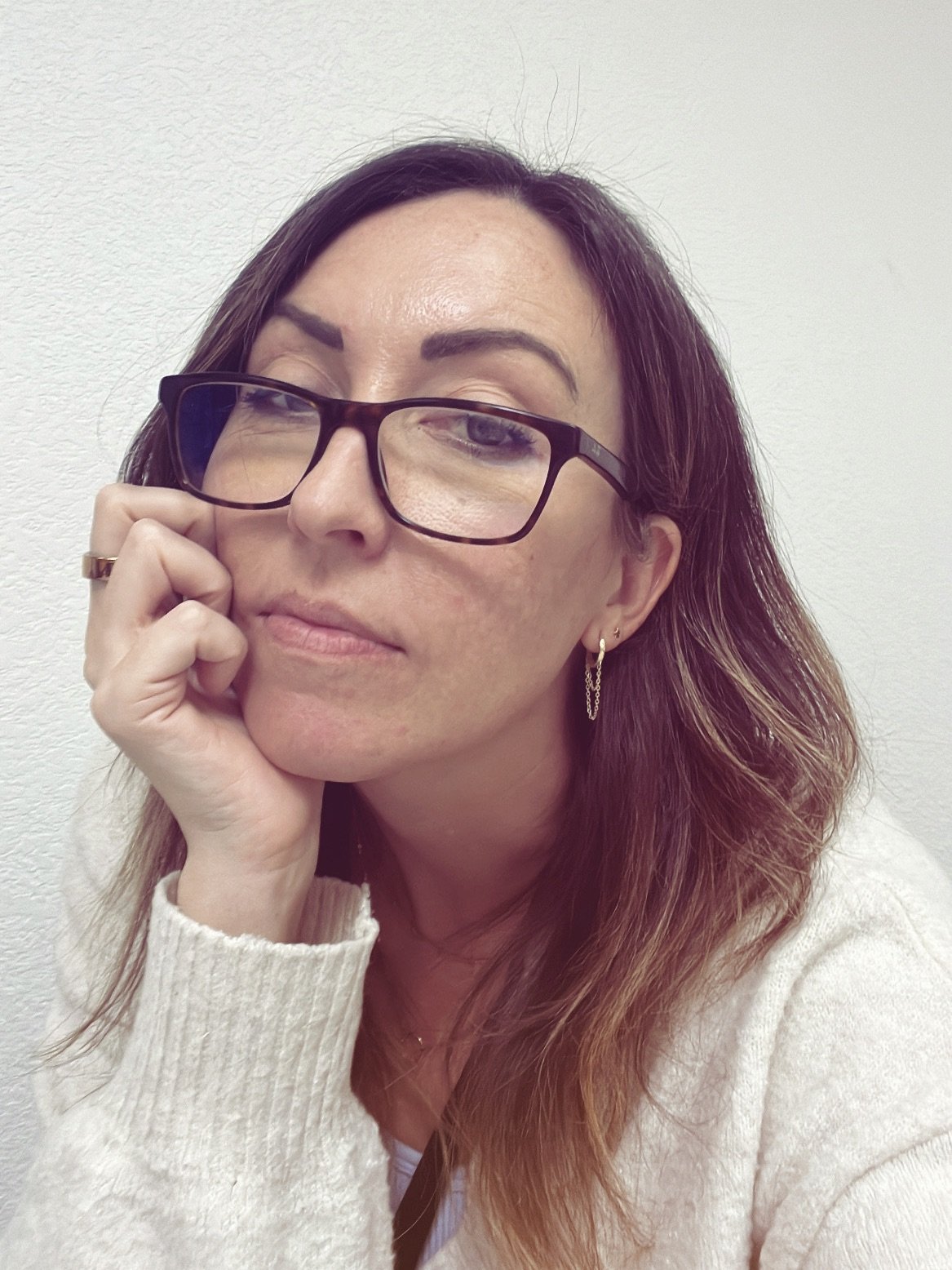 A woman with glasses and earrings resting her chin on her hand, looking at the camera with a neutral expression, against a plain white background.