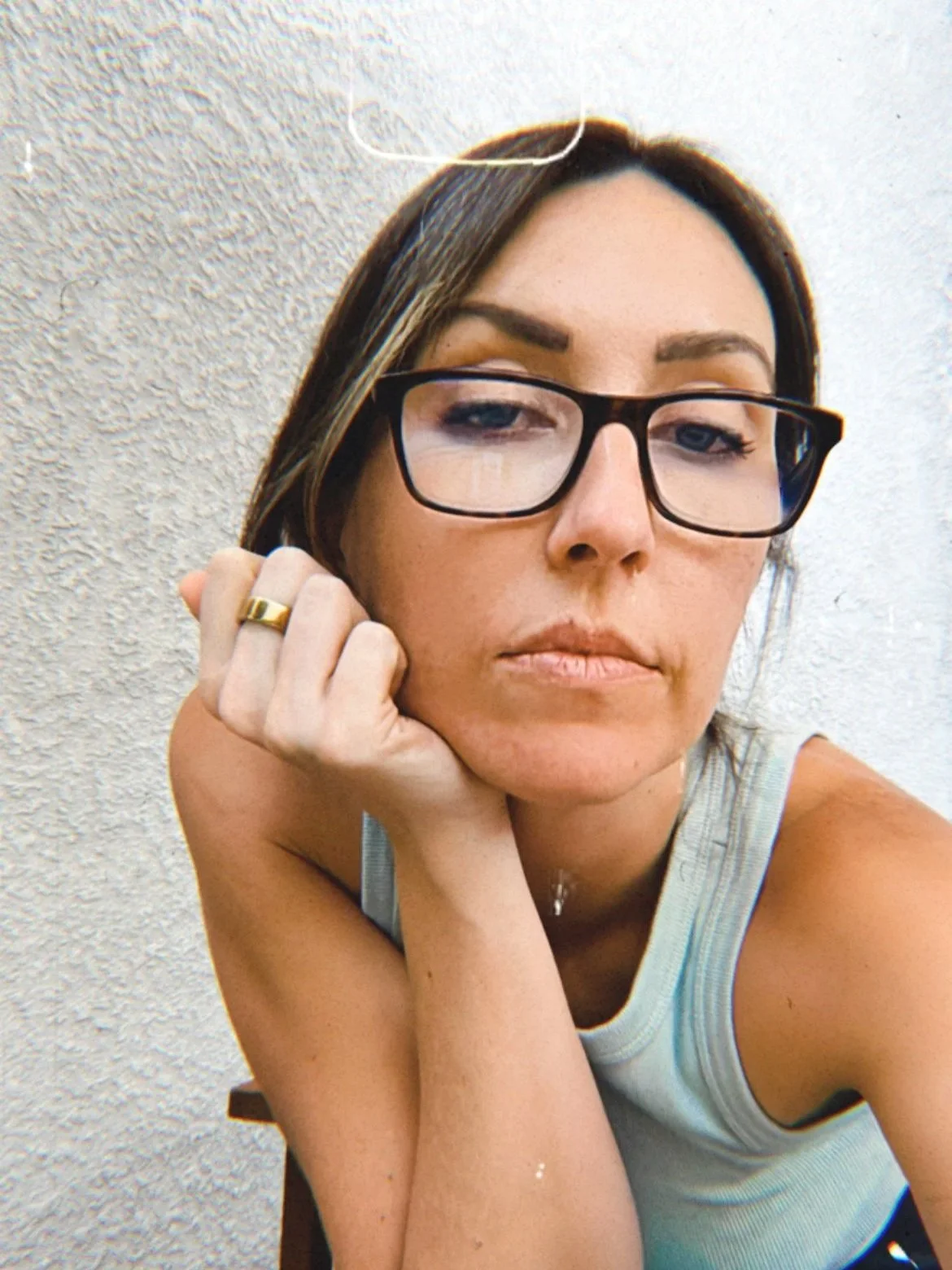 A young woman with brown hair, wearing glasses and a sleeveless top, resting her chin on her hand against a textured wall.