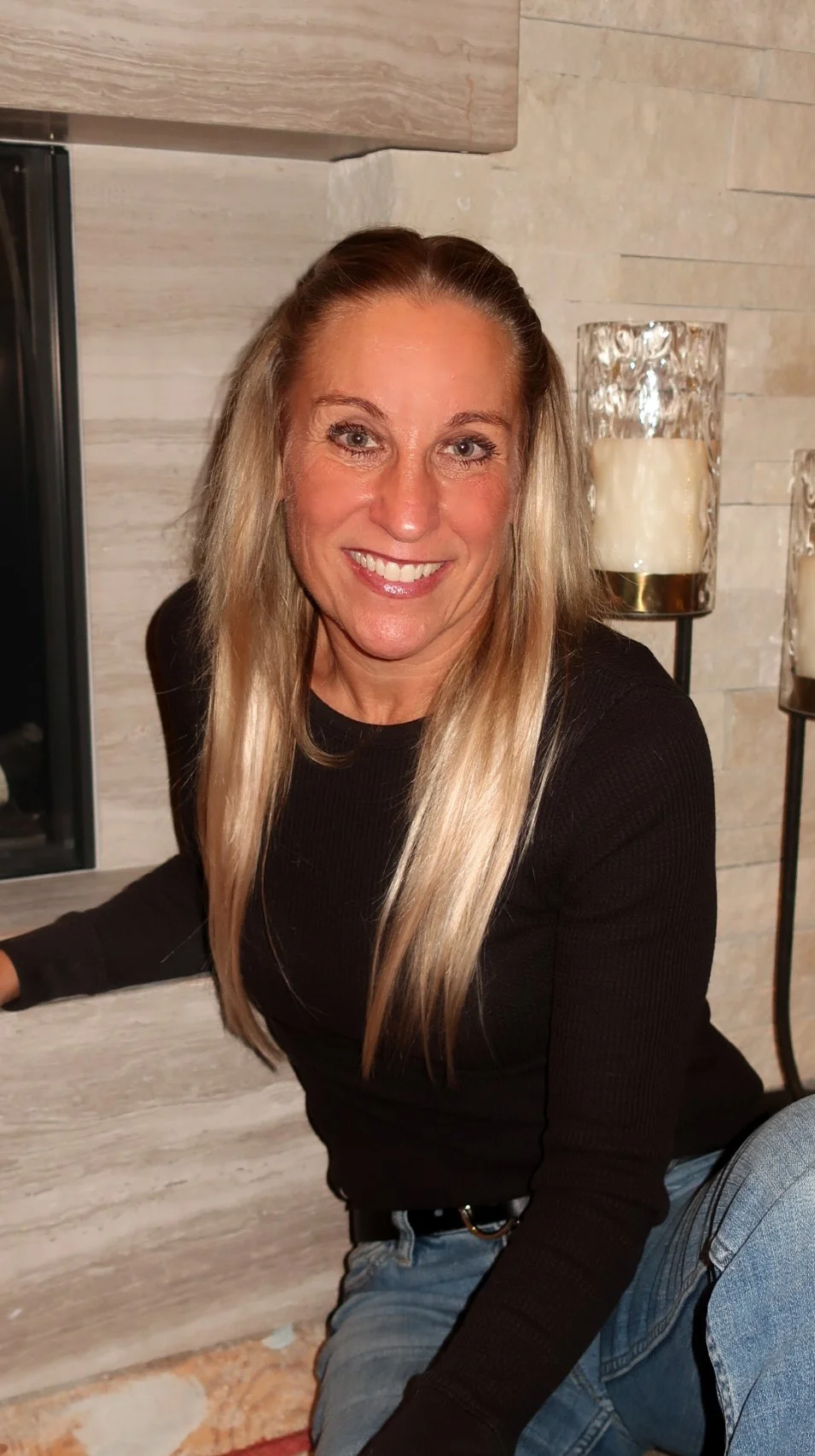 A woman with long blonde hair smiling, wearing a black long-sleeve shirt and blue jeans, sitting indoors near a fireplace with candles in glass holders.