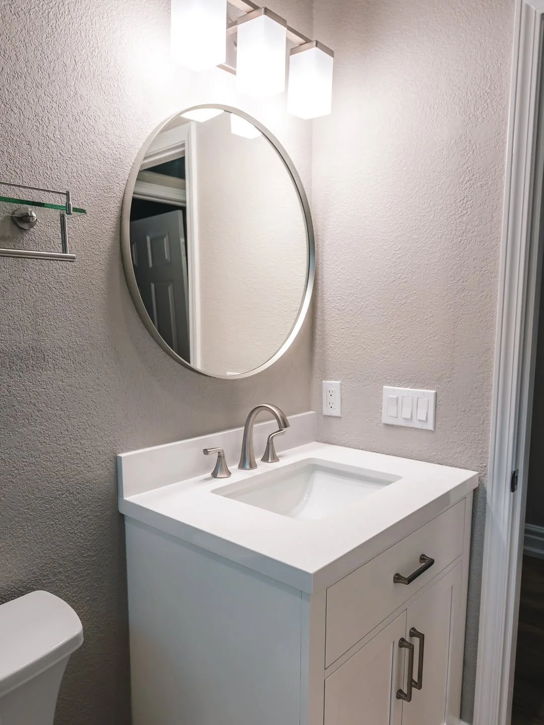 Bathroom vanity with white countertop, silver faucet, oval mirror, wall-mounted light fixture, electrical outlets, and a small towel rack on textured wall.