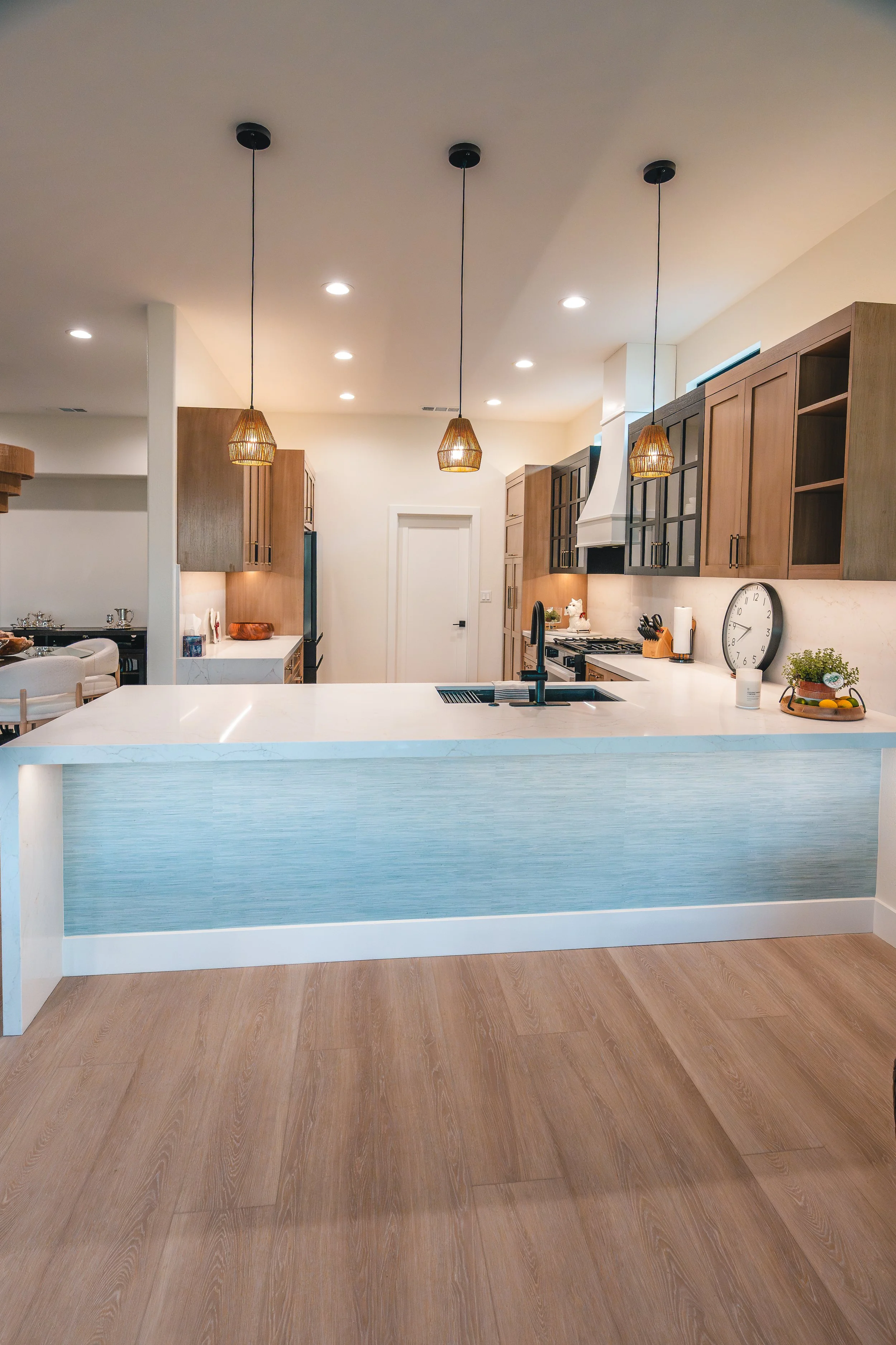 Modern kitchen with white island, wooden cabinets, pendant lighting, hardwood floors, clock, and some plants.