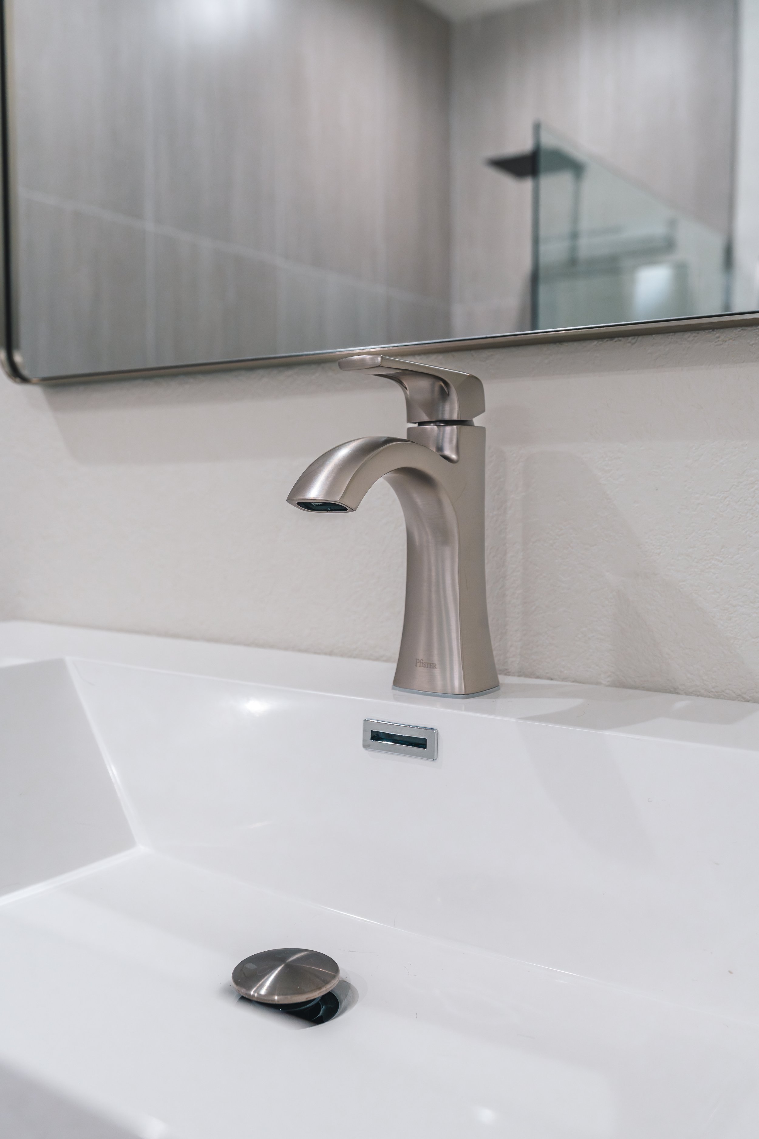 Modern bathroom sink with a brushed nickel faucet, white ceramic basin, and a mirror above, reflected in a tiled shower area.