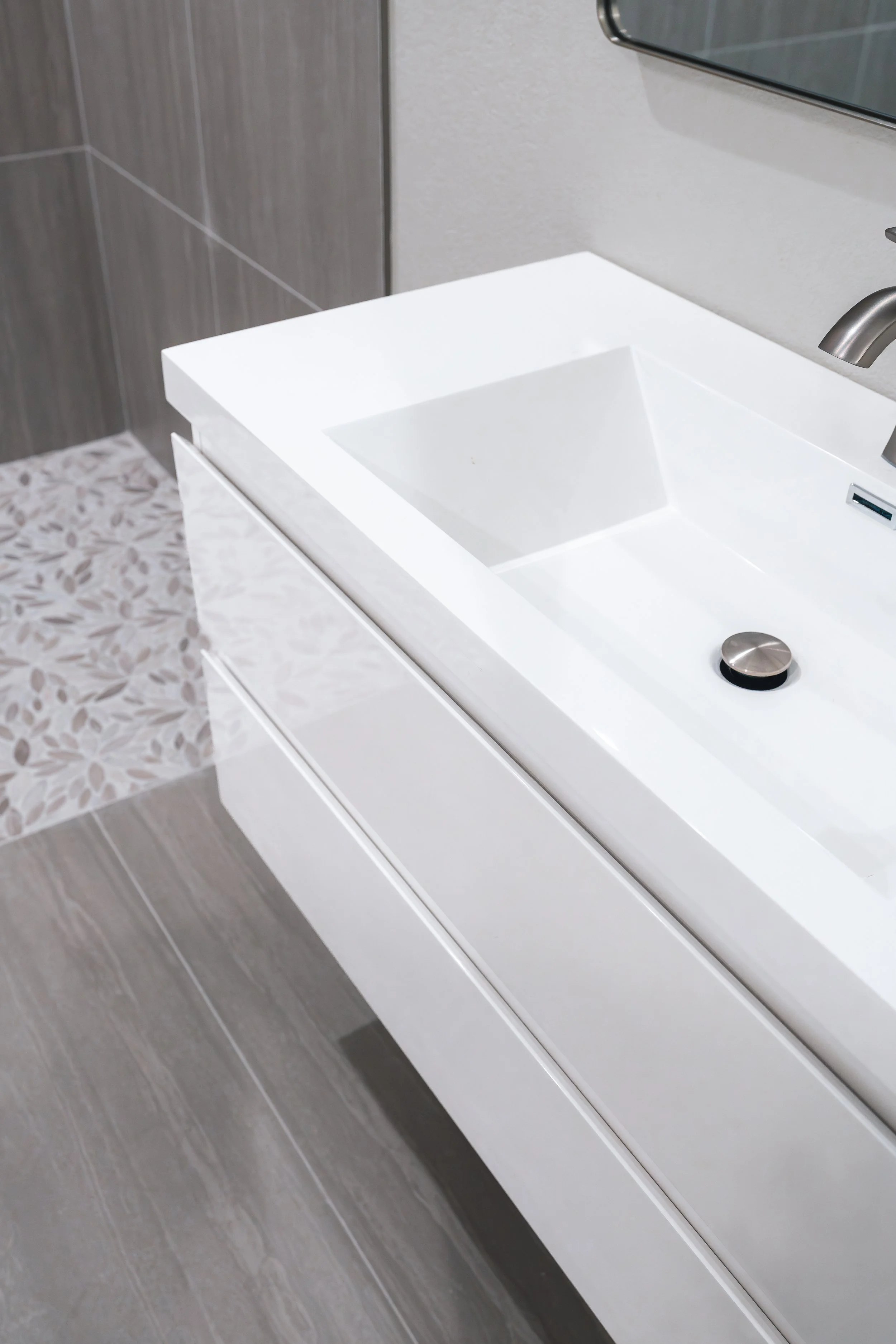 Close-up of a modern white bathroom sink with a silver faucet, set in a white vanity with drawer handles, against a wall with gray tiles and a mirror above.