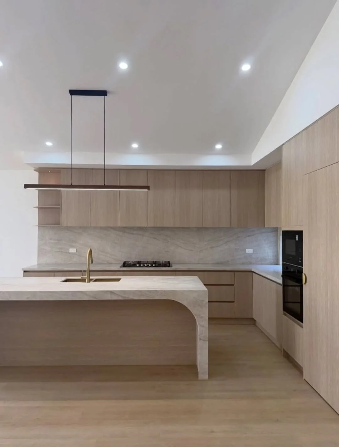 Modern kitchen with light wood cabinetry, marble backsplash, island with built-in sink and faucet, built-in oven and microwave, and recessed ceiling lights.
