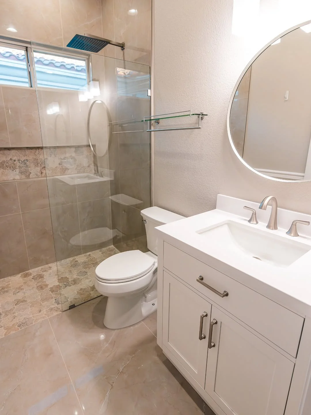Modern bathroom with a white vanity, sink, and oval mirror. Behind a glass partition is a walk-in shower with a rainfall showerhead. There is a toilet next to the vanity and beige tiled walls and floor.