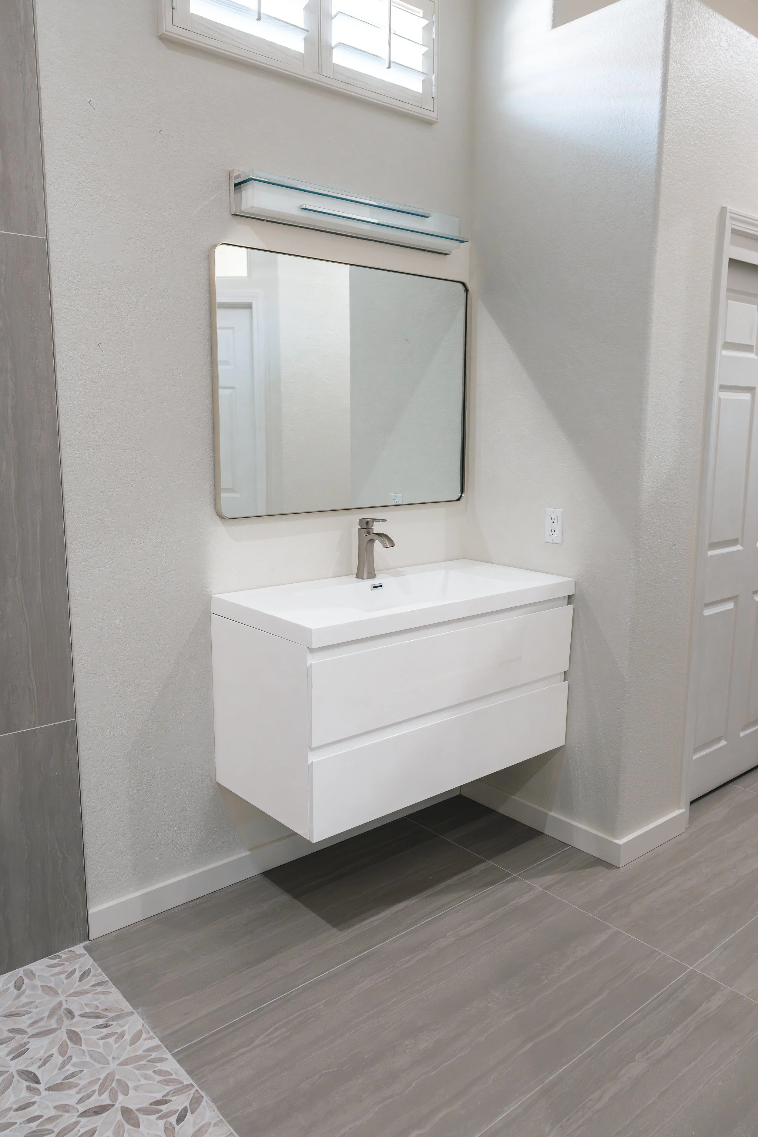 Modern white bathroom vanity with a rectangular mirror and a wall-mounted light fixture, with gray tiled flooring and a small window above.