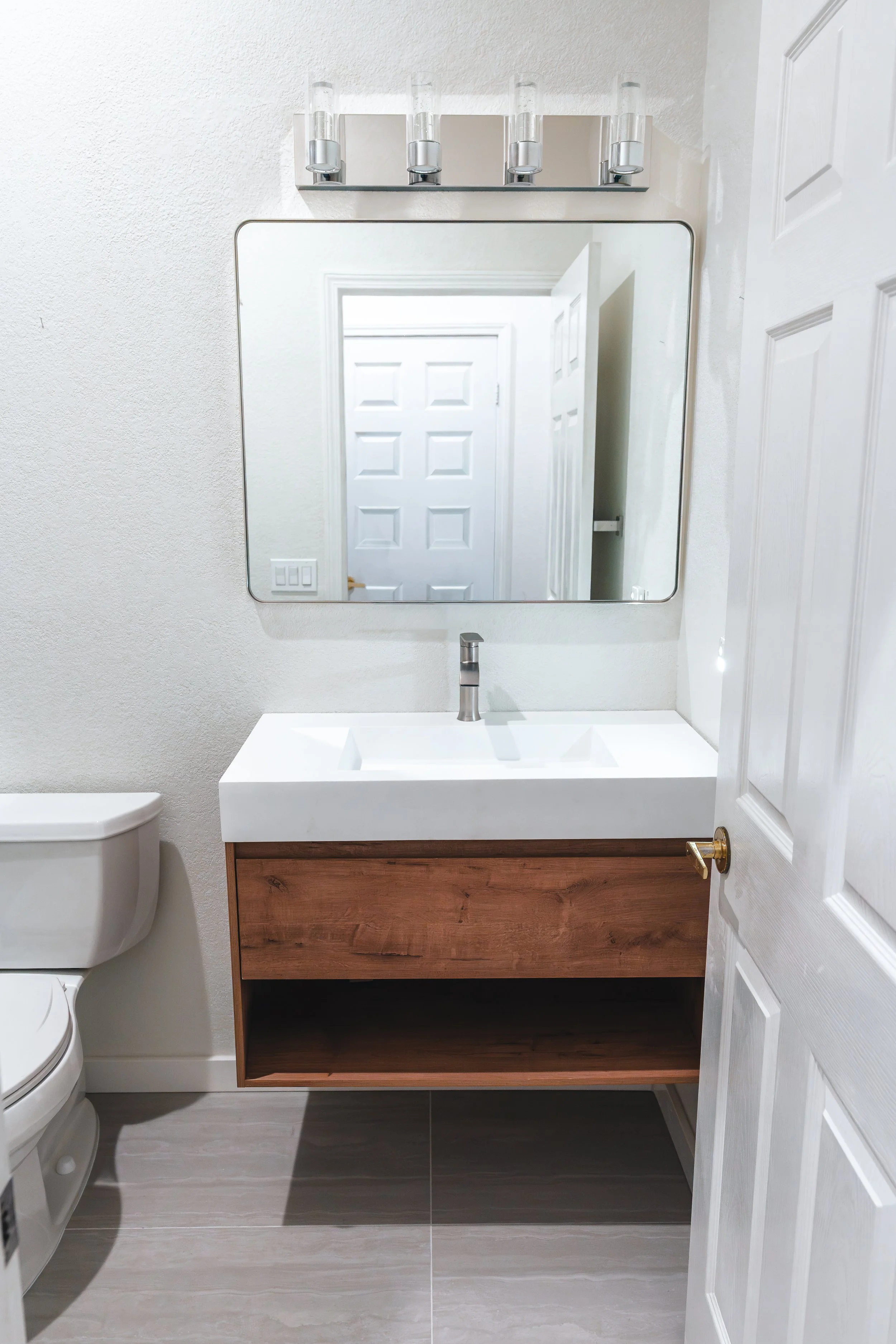 Bathroom with a white wall, a rectangular mirror, a modern wooden vanity with an open shelf, a white sink with a silver faucet, a white toilet, and a light fixture with four glass shades above the mirror.