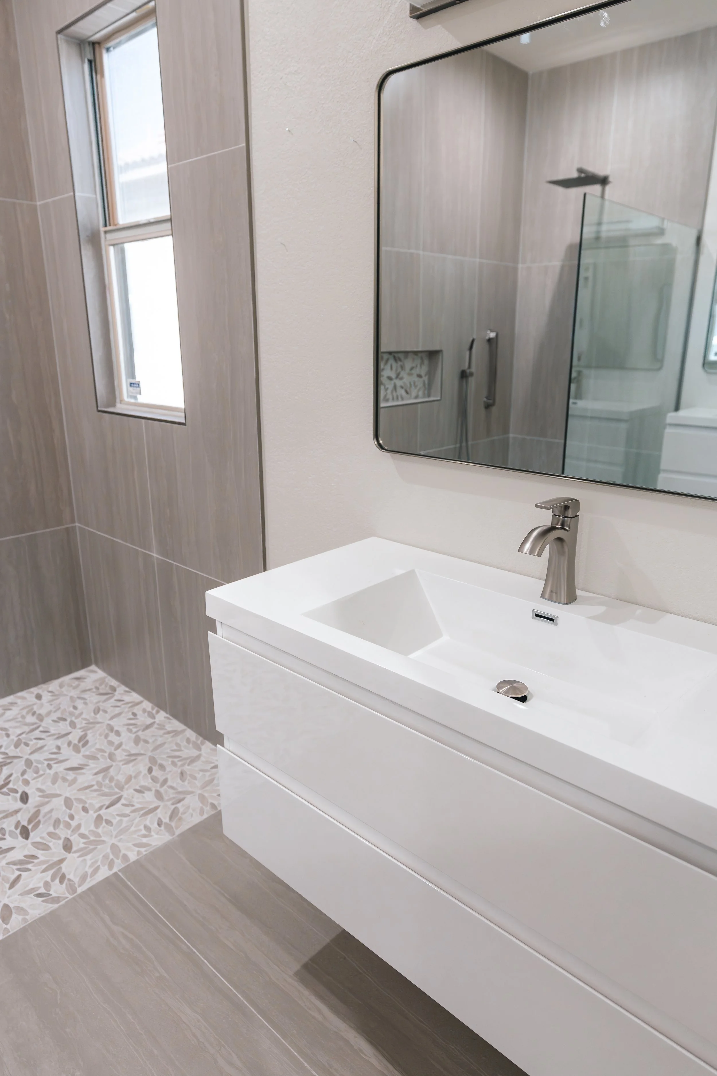 Modern bathroom with beige tile walls, a floating white vanity with a built-in sink, a large mirror, a window, and a walk-in shower with a glass enclosure and rainfall showerhead.