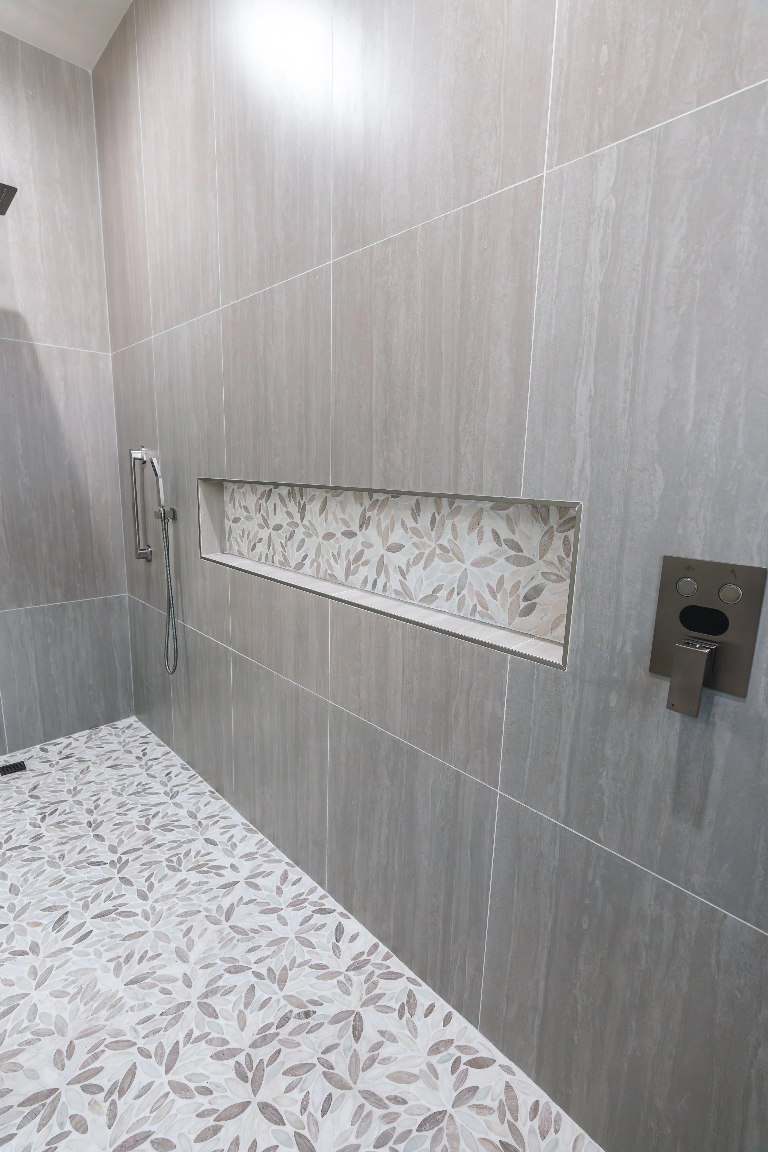 A modern shower with beige and gray tiles, a built-in niche with leaf-patterned tiles, a handheld showerhead, and a digital control panel.