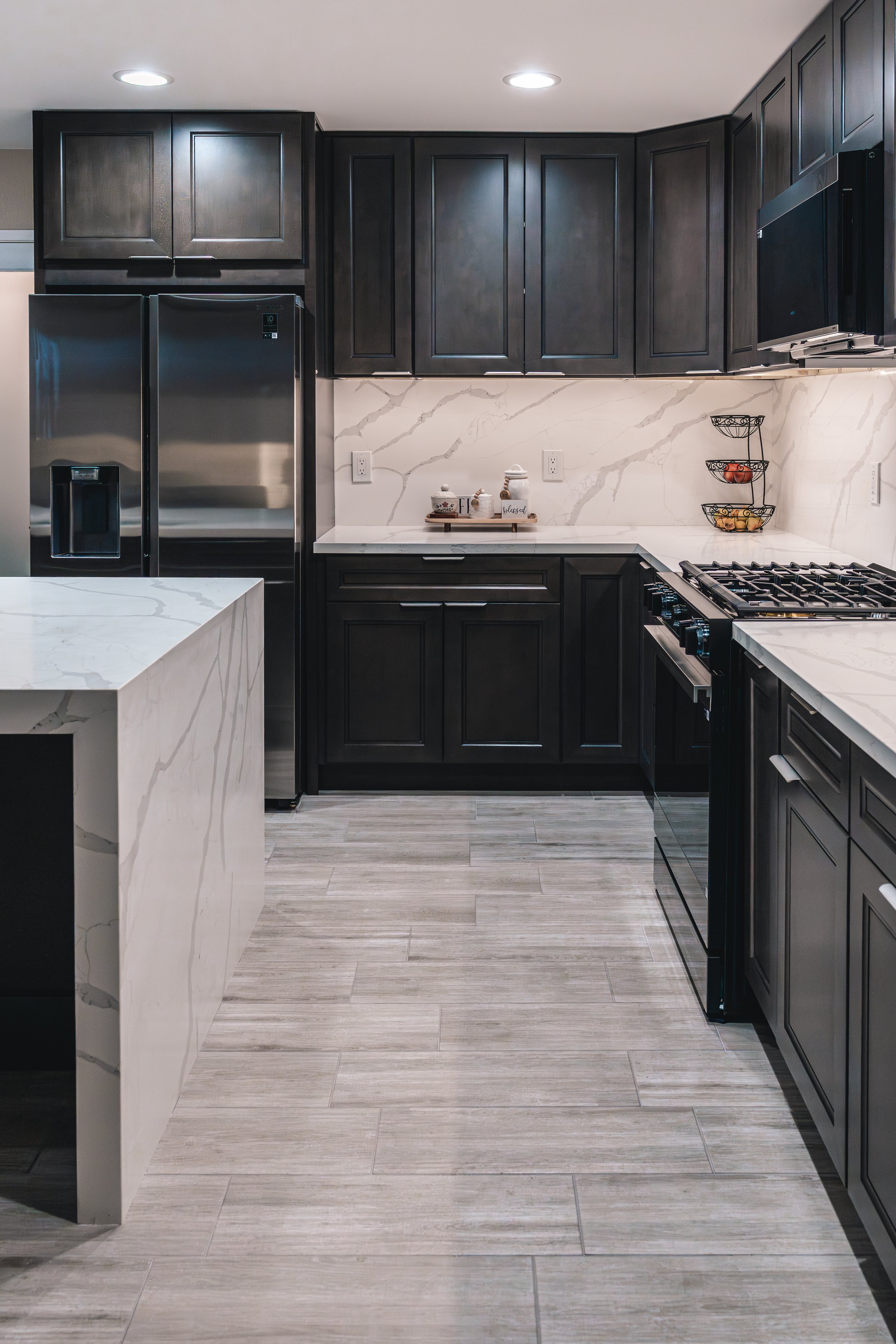 Modern kitchen with black cabinets, stainless steel refrigerator, marble backsplash, countertop, and built-in oven with gas stove.
