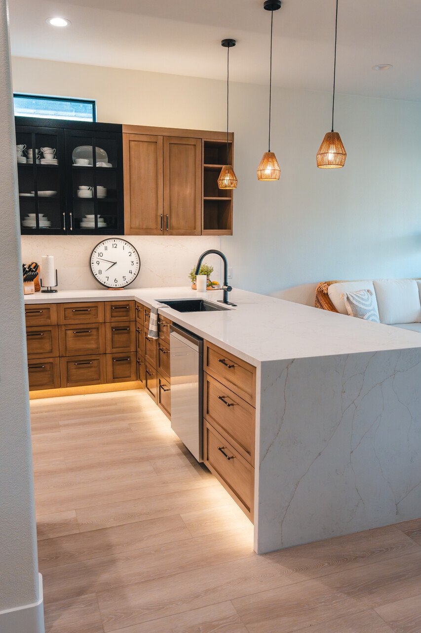 Modern kitchen with wooden cabinets, white marble countertops, black sink faucet, pendant lights, a wall clock, and a cozy living space with a white couch.