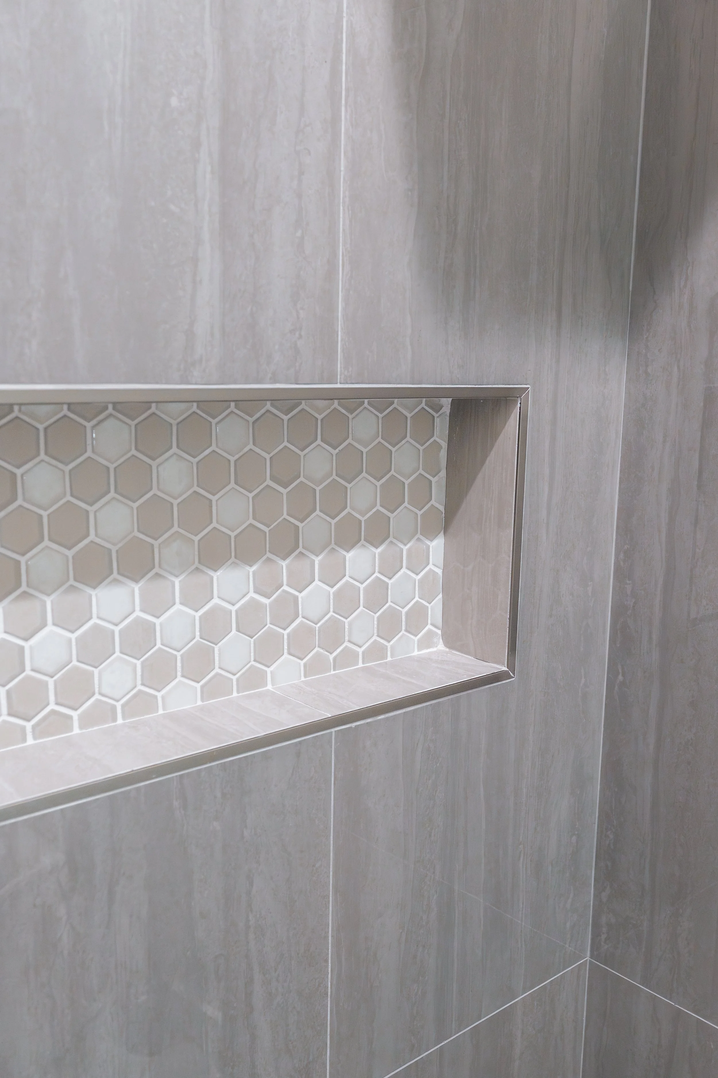 Close-up of a recessed shower shelf with beige hexagonal tile mosaic, surrounded by large gray wall tiles.